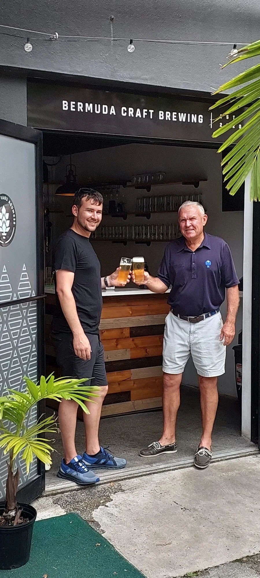 Two men standing at the entrance of Bermuda Craft Brewing, toasting with glasses of beer inside the brewery.