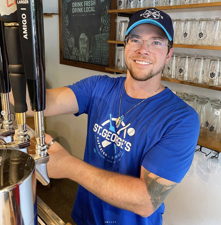 A young man with glasses and a tattoo on his arm, wearing a blue St. George's T-shirt and cap, pouring beer from a tap at a bar or brewery.