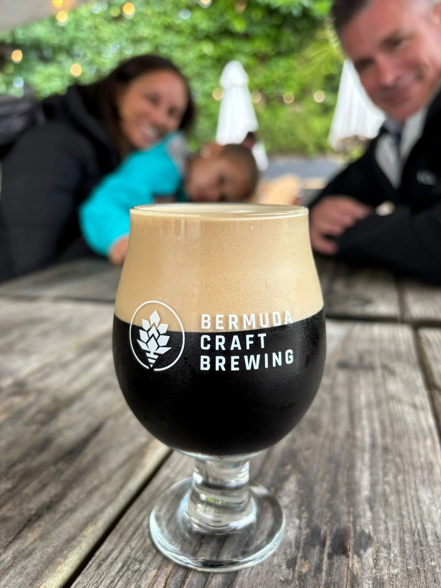 A glass of dark stout beer with a foamy head on a wooden table, with a blurred family in the background.