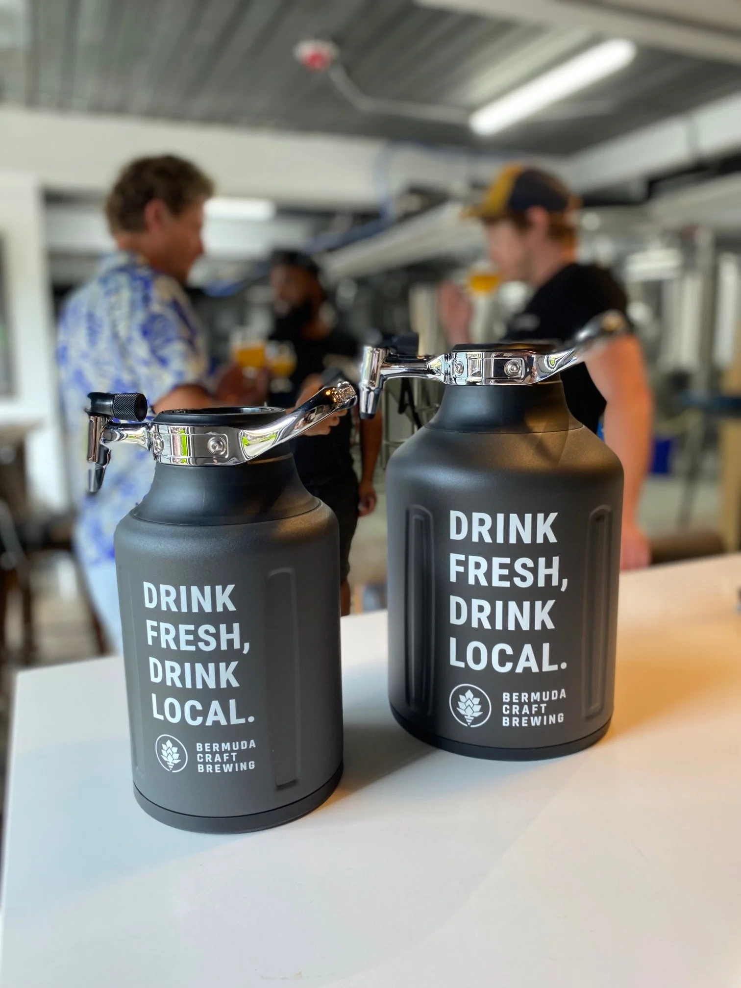 Two black growlers with silver handles and spouts on a white table, labeled with the slogan 'Drink Fresh, Drink Local' and the logo for Bermuda Craft Brewing. In the background, three people are socializing, holding drinks, and the environment appears to be a brewery or taproom.