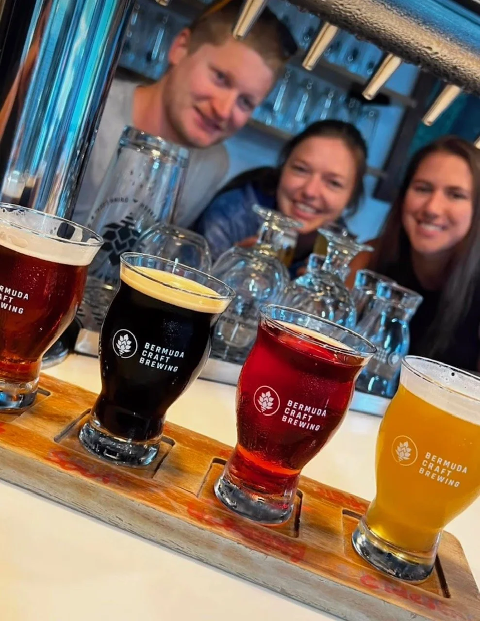 Three people smiling at a bar with a flight of four different beers in front of them, each beer in a glass with 'Bermuda Craft Brewing' logo, and glasses and bottles in the background.