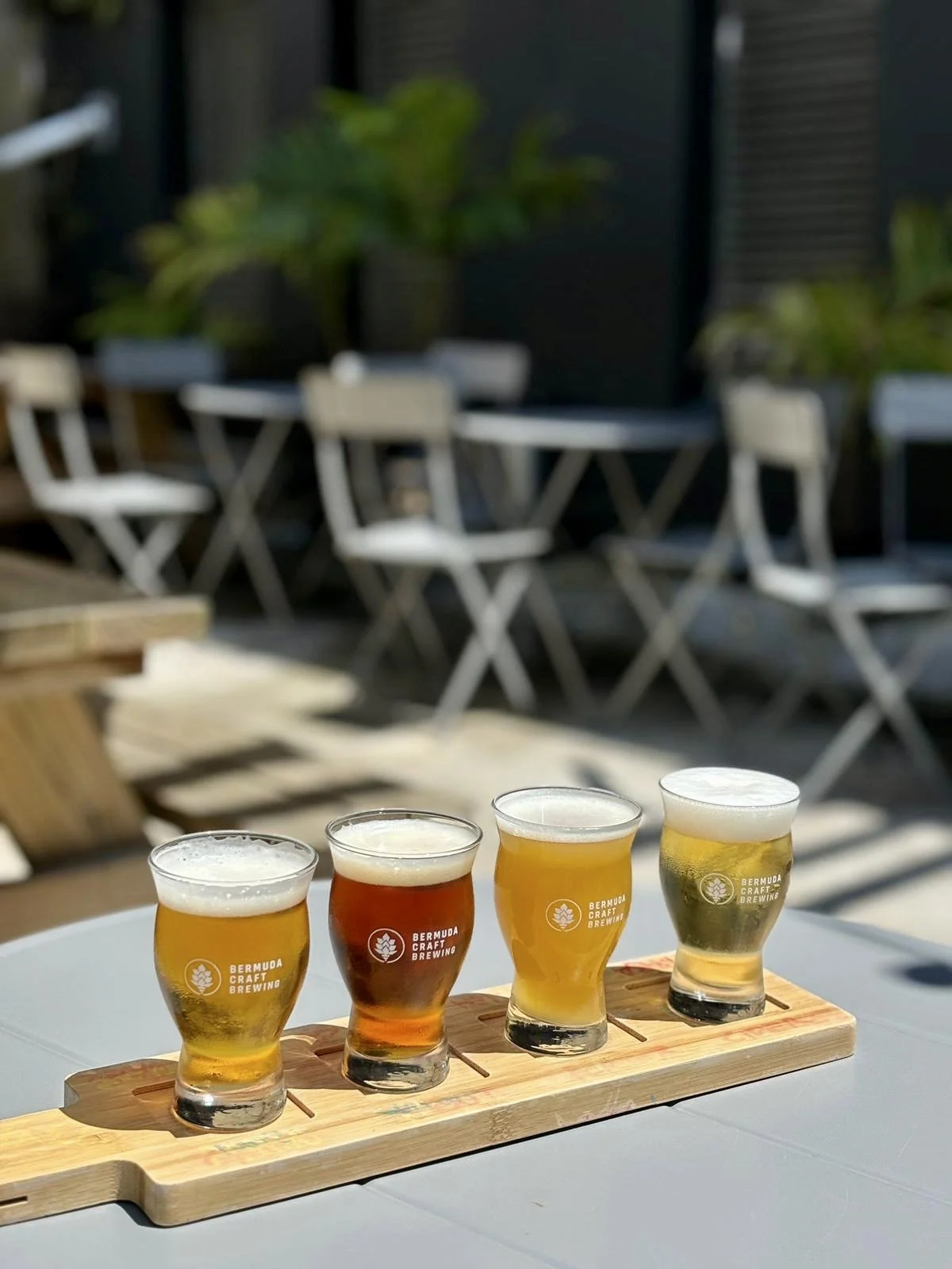 A flight of four different beers served in small glasses, placed on a wooden flight paddle on an outdoor table.