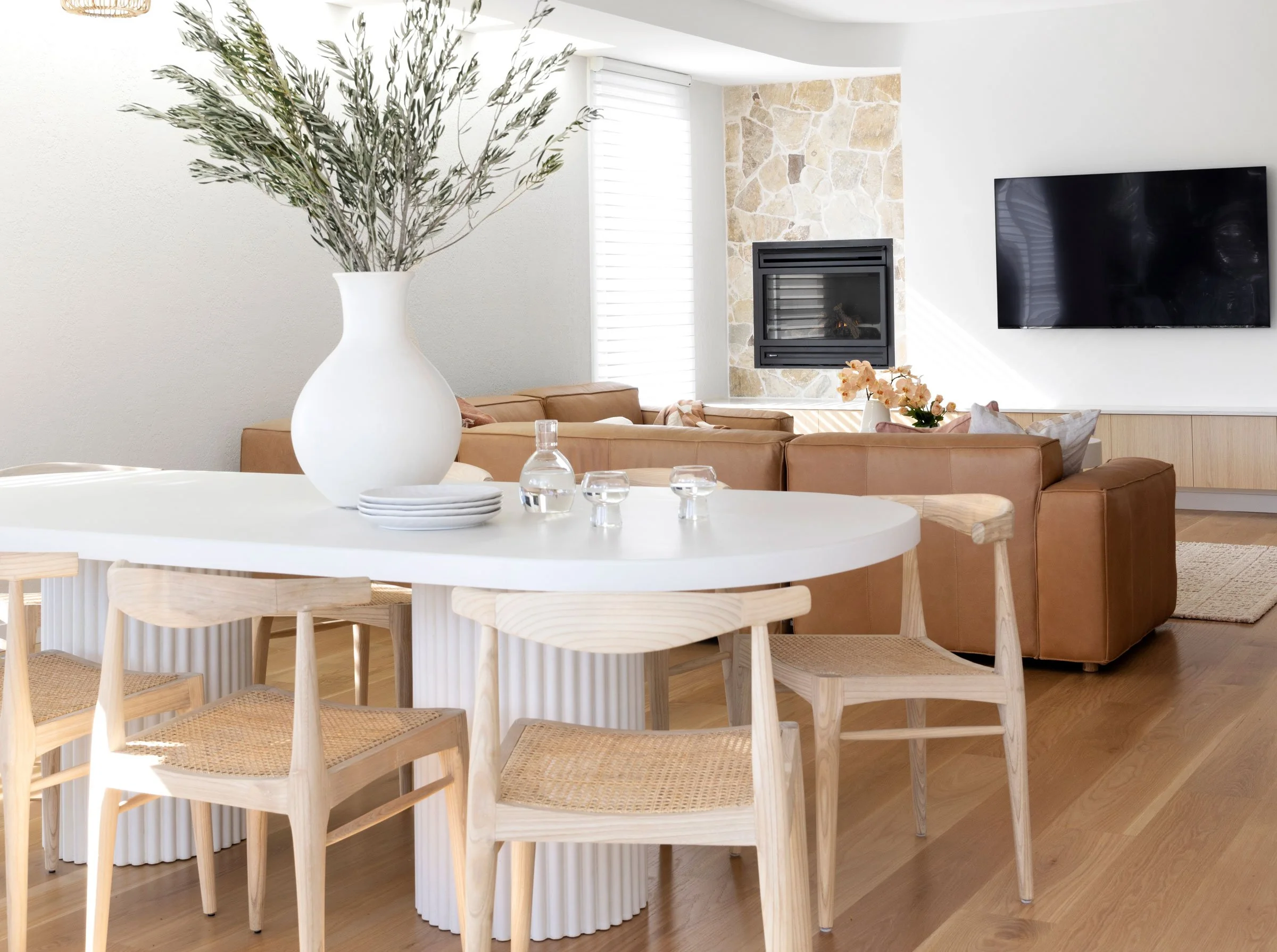 Modern living room with a flat-screen TV, a beige sectional sofa with floral pillows, a stone fireplace, white walls, and a large floral arrangement in a white vase on a dining table.