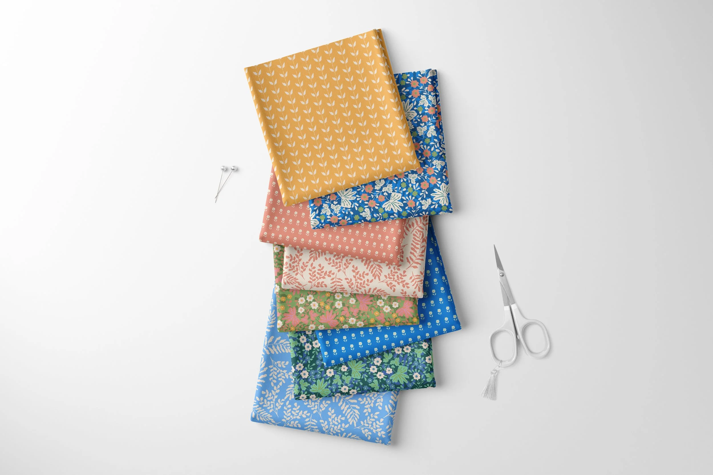 A stack of colorful patterned fabric squares with floral and geometric designs on a white background, next to sewing pins and scissors. Fabrics designed by Nelly and Isobel.