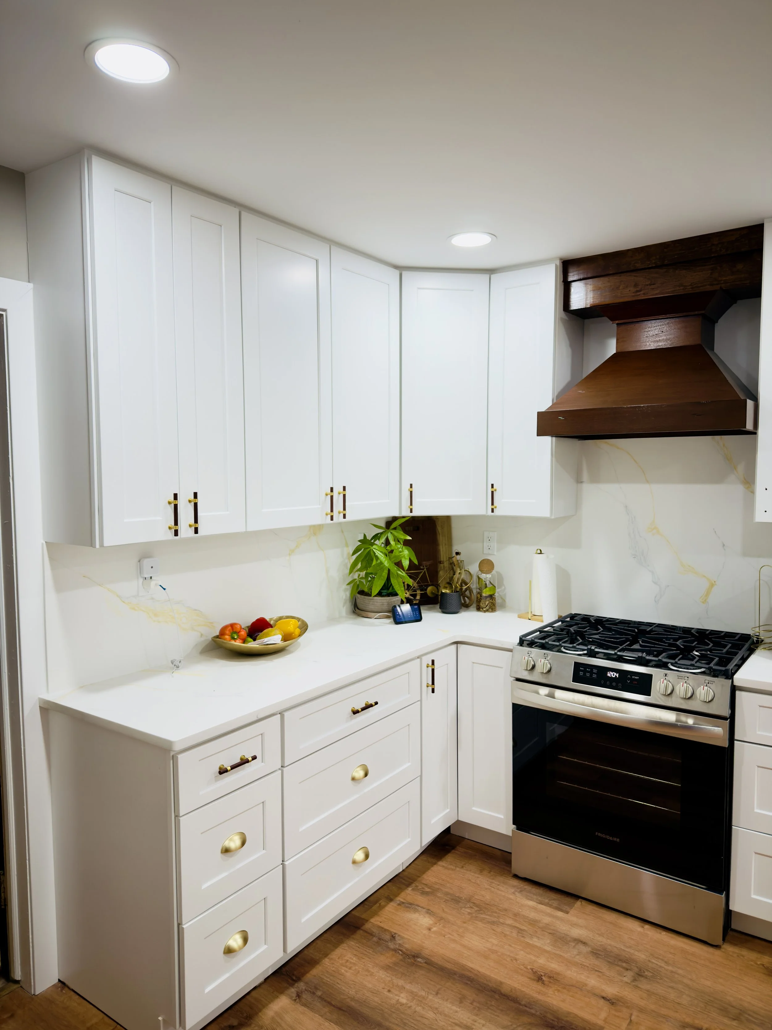 A kitchen with white cabinets, gold handles, a wooden range hood, a stove, and a marble backsplash. There is a plant, a bowl of vegetables, and kitchen accessories on the countertop.