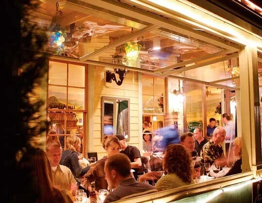 People dining inside a cozy restaurant with large windows and warm lighting, visible through an open window.