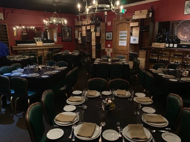 A dimly lit restaurant dining room with round tables set with plates, glasses, beige napkins, and cutlery. The room has dark green chairs, wooden shelves with wine bottles, and red walls with framed pictures. Chandeliers hang from the ceiling.