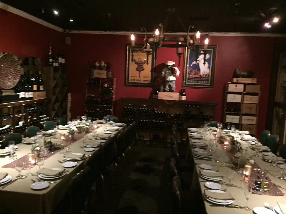 A dimly lit restaurant or private dining room with a long table set for a meal, wine bottles on shelves, and framed artwork on a dark red wall, with a chandelier overhead.