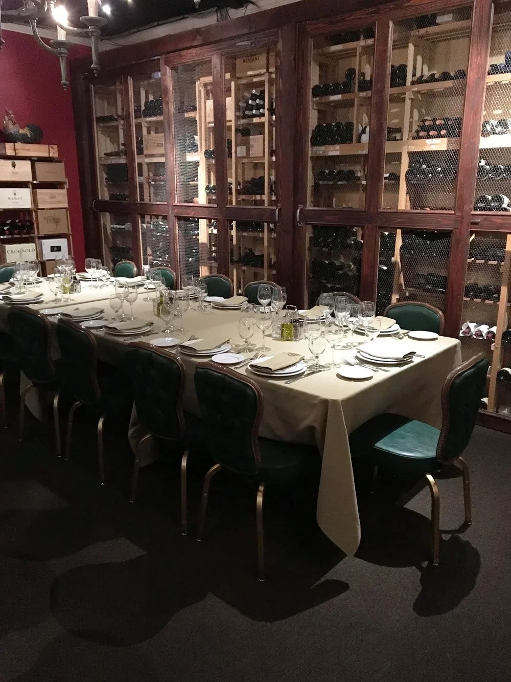 A long dining table set with plates, glasses, and napkins in a wine cellar with wooden shelves filled with wine bottles behind a glass partition.
