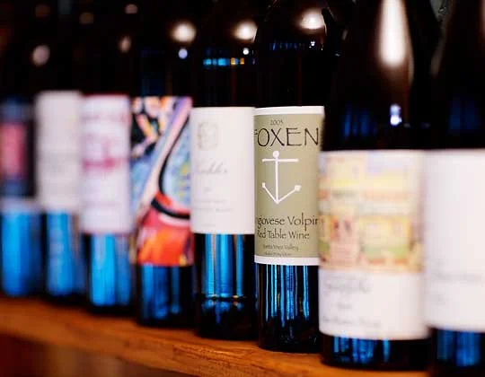 A row of wine bottles on a wooden shelf, with labels facing forward, including one labeled 'Oxygen' with an anchor design.