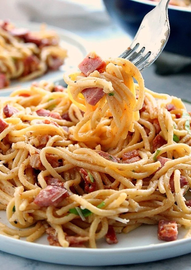 Close-up of a fork lifting a bite of spaghetti with bits of bacon and green herbs on a white plate.