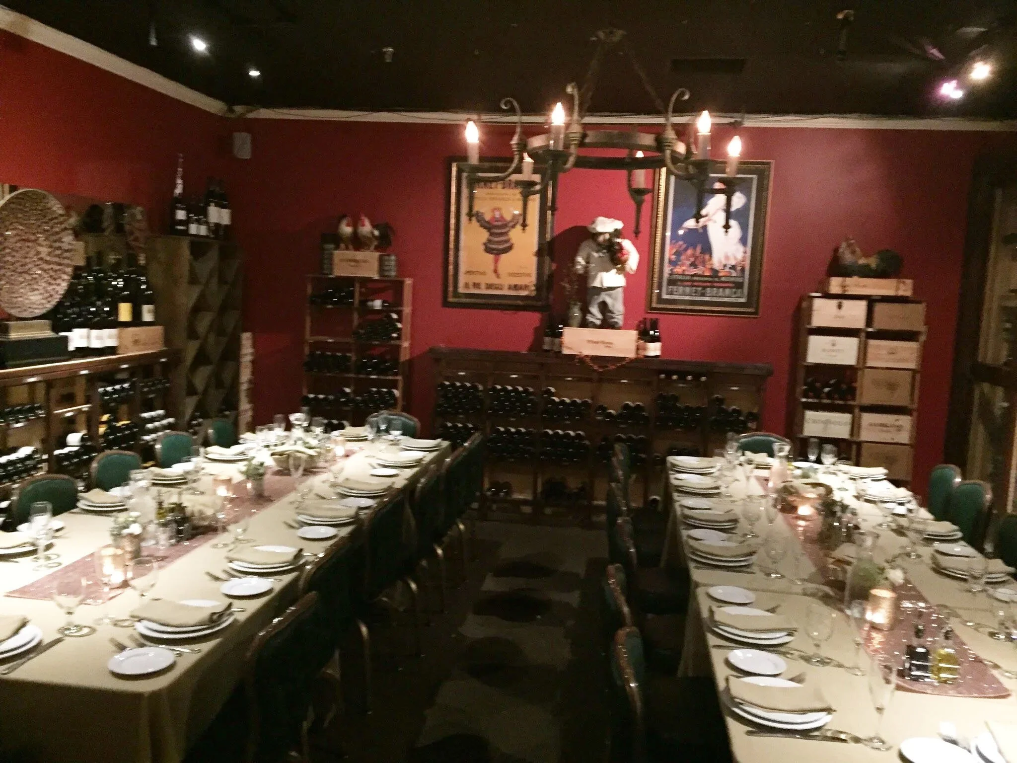 A restaurant dining room decorated for a formal event, with tables set with white tablecloths, plates, glasses, and silverware, wine bottles on shelves, and a black chandelier hanging from the ceiling, featuring vintage movie posters on the walls.