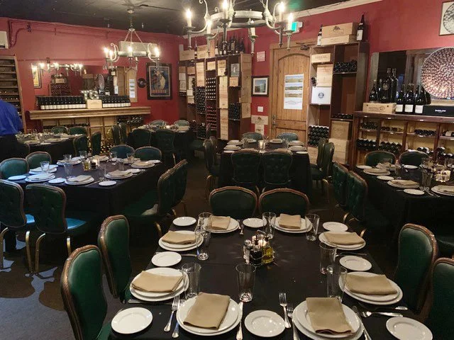 A cozy restaurant with round tables set with place settings, green cushioned chairs, wine bottles on shelves, and warm lighting from chandeliers.