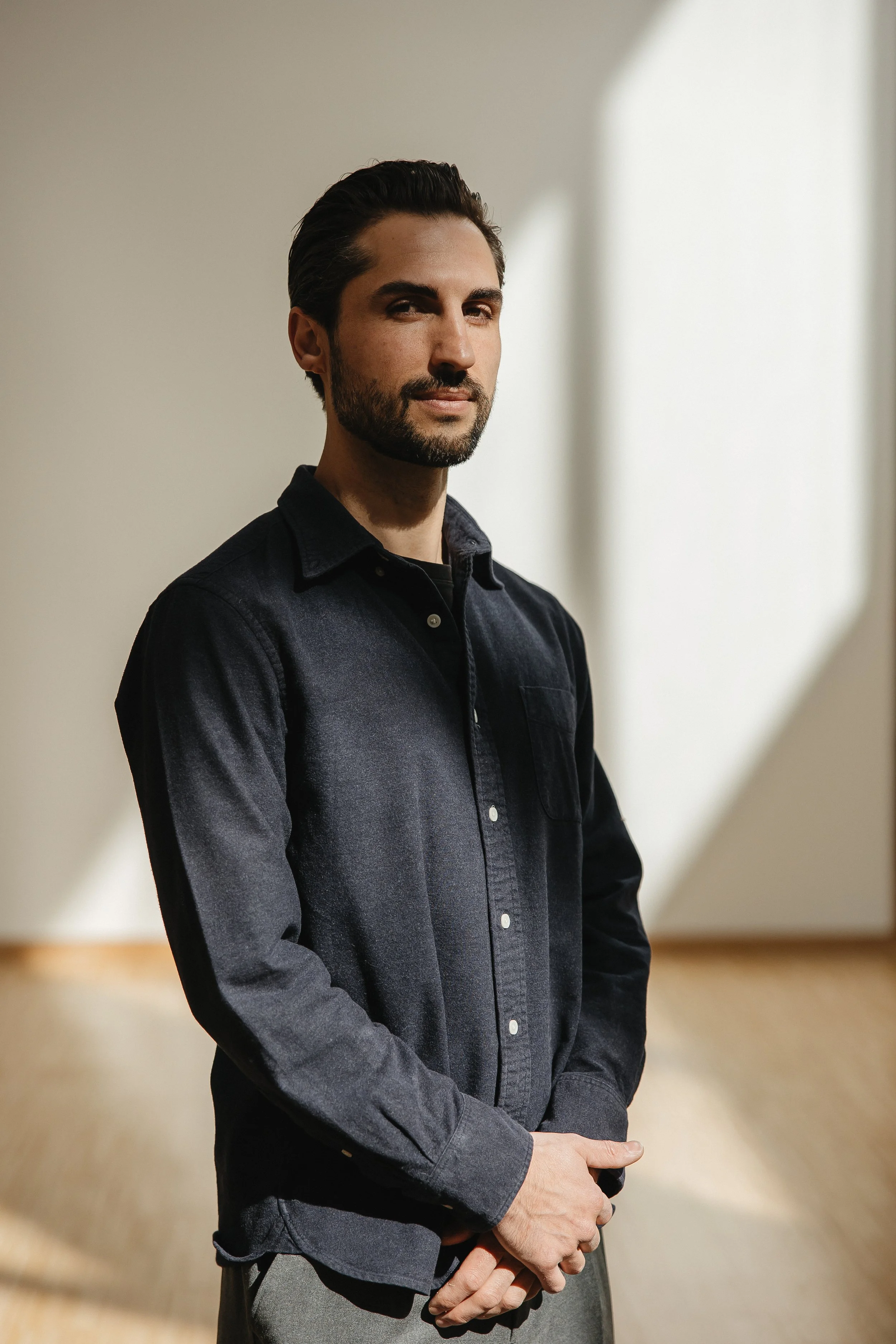 Un homme avec des cheveux bruns et une barbe, portant une chemise noire, se tient dans une pièce bien éclairée avec un fond blanc et un sol en bois.
