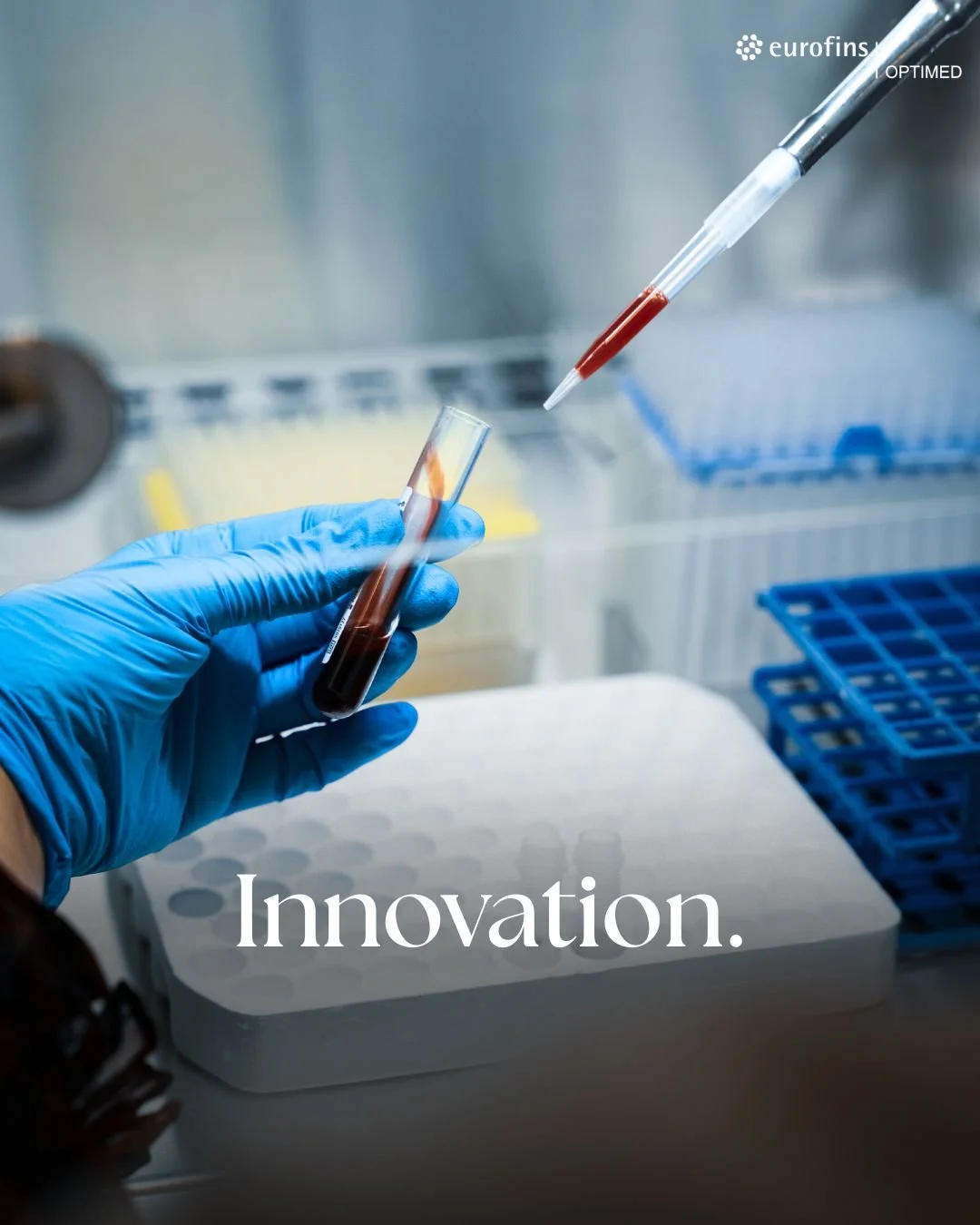 Une main gantée de bleu manipulant un tube à essai contenant un liquide rouge, dans un laboratoire de recherche avec équipements scientifiques en arrière-plan.