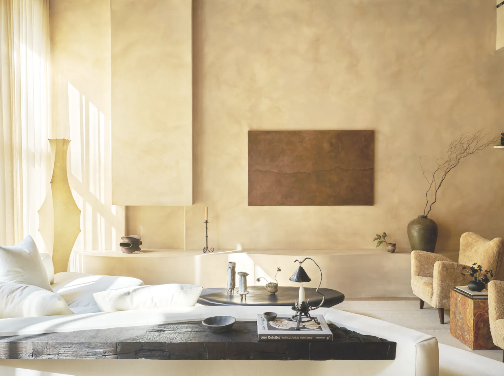 Salon moderne avec canapé blanc, table basse noire, tableau abstrait, fauteuil en peluche beige, vases décoratifs et grandes fenêtres laissant entrer la lumière naturelle.
