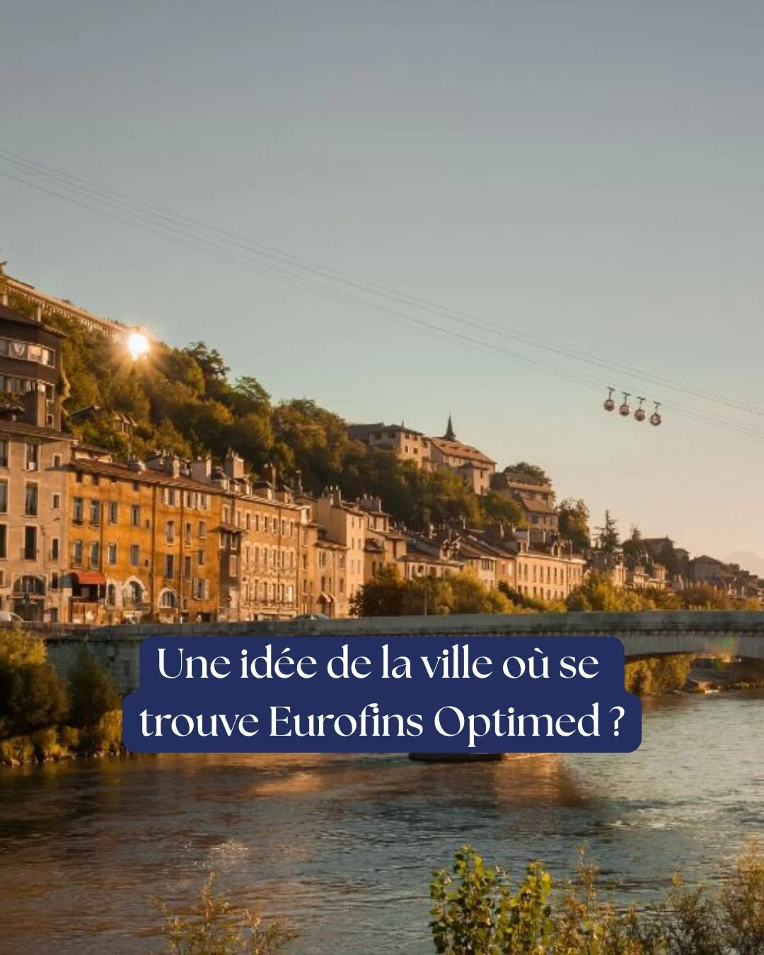 Une vue d'une ville construite sur une colline avec des bâtiments en pierre, un fleuve au premier plan et une télécabine en suspension dans le ciel clair.