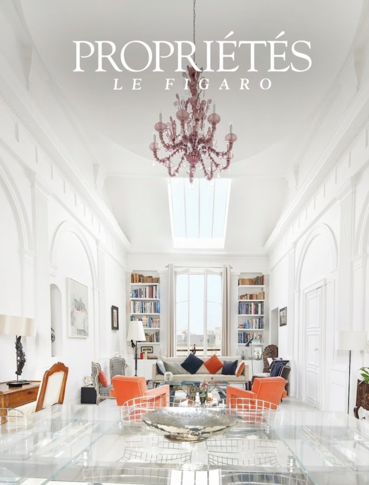 Salon lumineux avec mobilier moderne, livres sur étagères, chandelier rose, et grande fenêtre