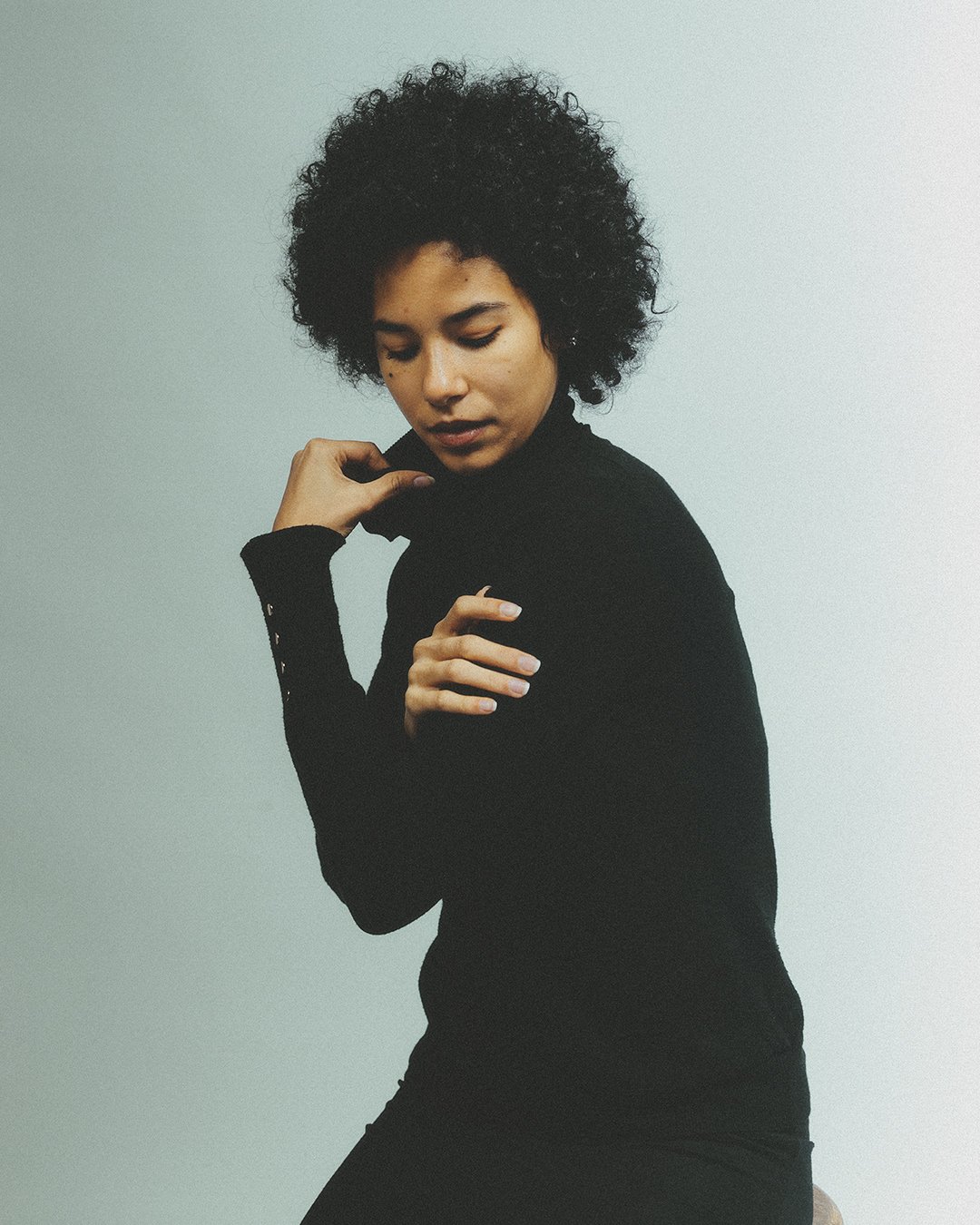 Femme portant un pull noir, cheveux bouclés, posant dans un studio avec fond neutre.