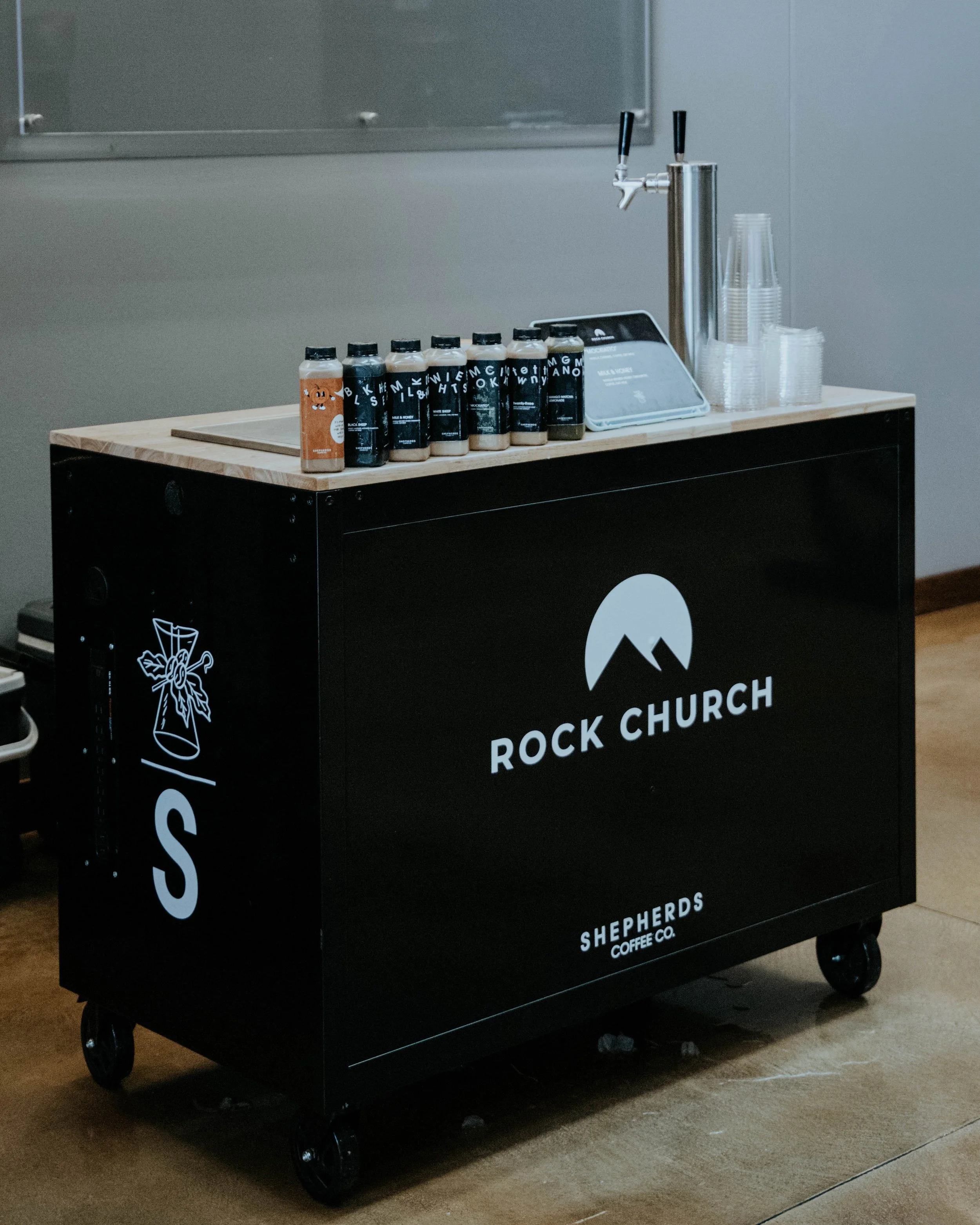 A mobile coffee cart with the logo 'Rock Church Shepherds Coffee Co.' on the front, featuring bottles of syrup, disposable cups, and a coffee tap on top.