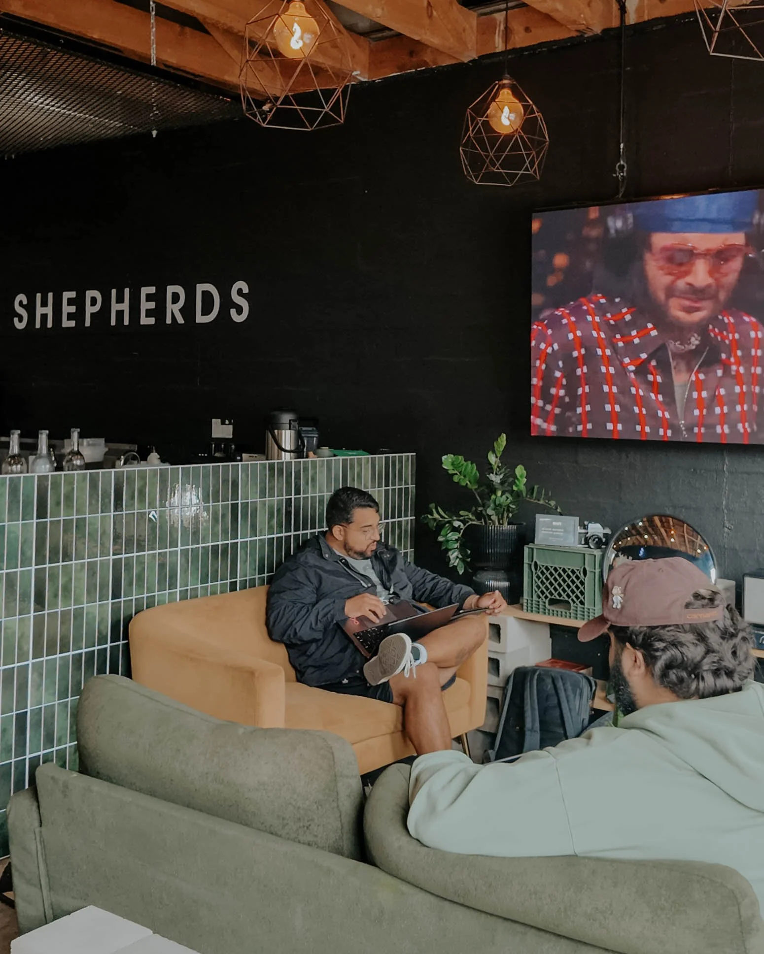 Two men sit on beige and green couches in a cafe or lounge with a black wall that has the word 'SHEPHERDS' in white letters. One man is using a laptop, while the other, wearing a cap, is facing him. A large screen showing a person in a red checkered shirt is mounted on the wall. Decorative lights hang from the ceiling, and there are green plants and miscellaneous items on a shelf near the wall.