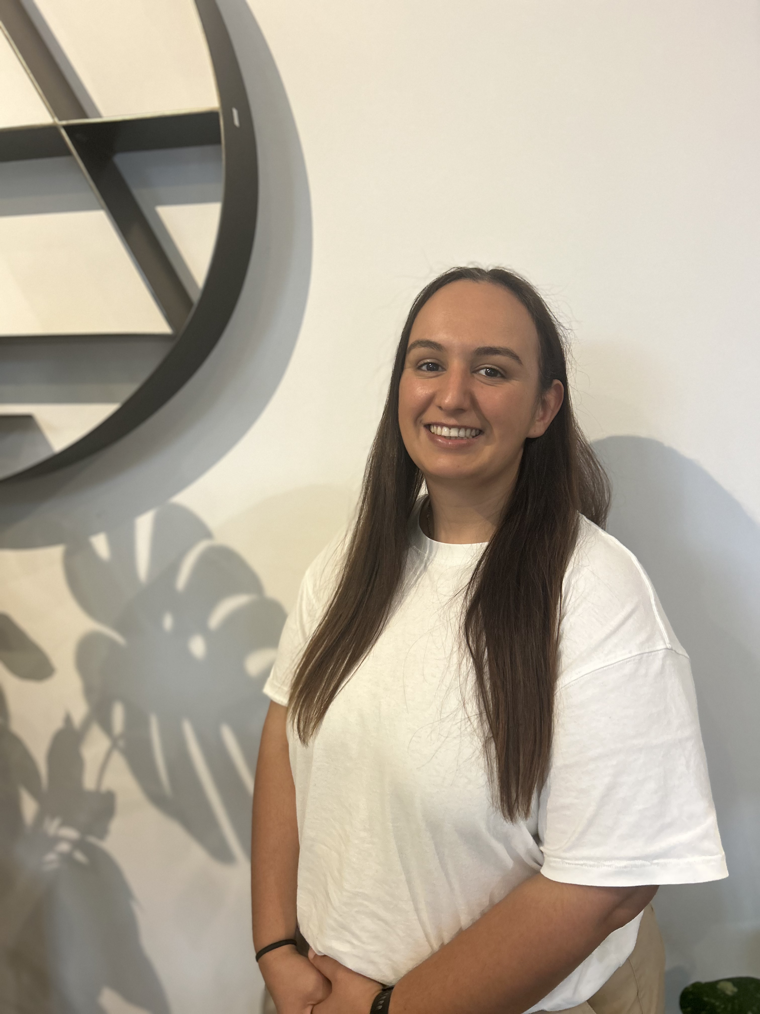 A young woman with light brown hair and blue eyes, smiling, wearing a white t-shirt layered with a light gray jacket and black pants, standing against a white background.