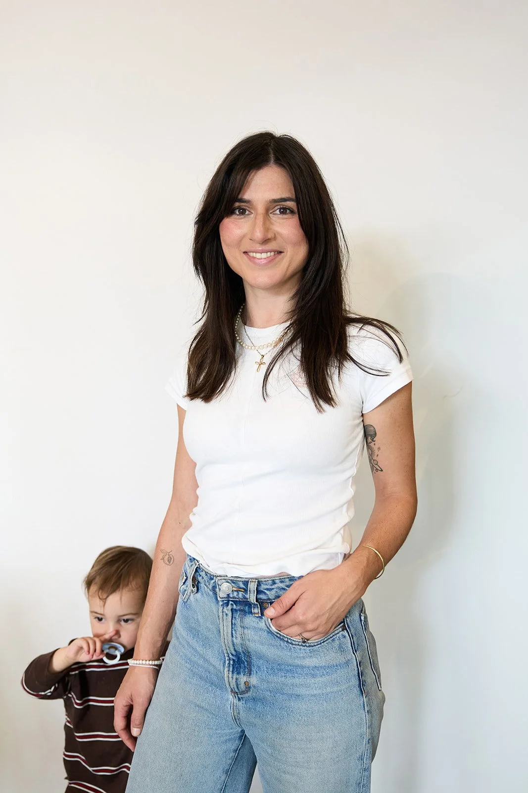 A smiling woman with wavy dark hair and a flower tattoo on her arm, wearing a sleeveless light-colored top and black pants, standing against a plain white background.