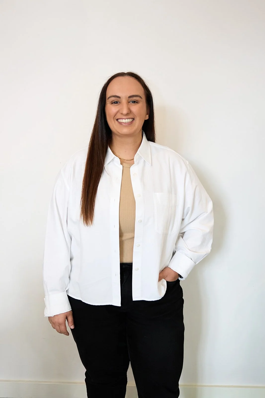 A young woman with light brown hair and blue eyes, smiling, wearing a white t-shirt layered with a light gray jacket and black pants, standing against a white background.
