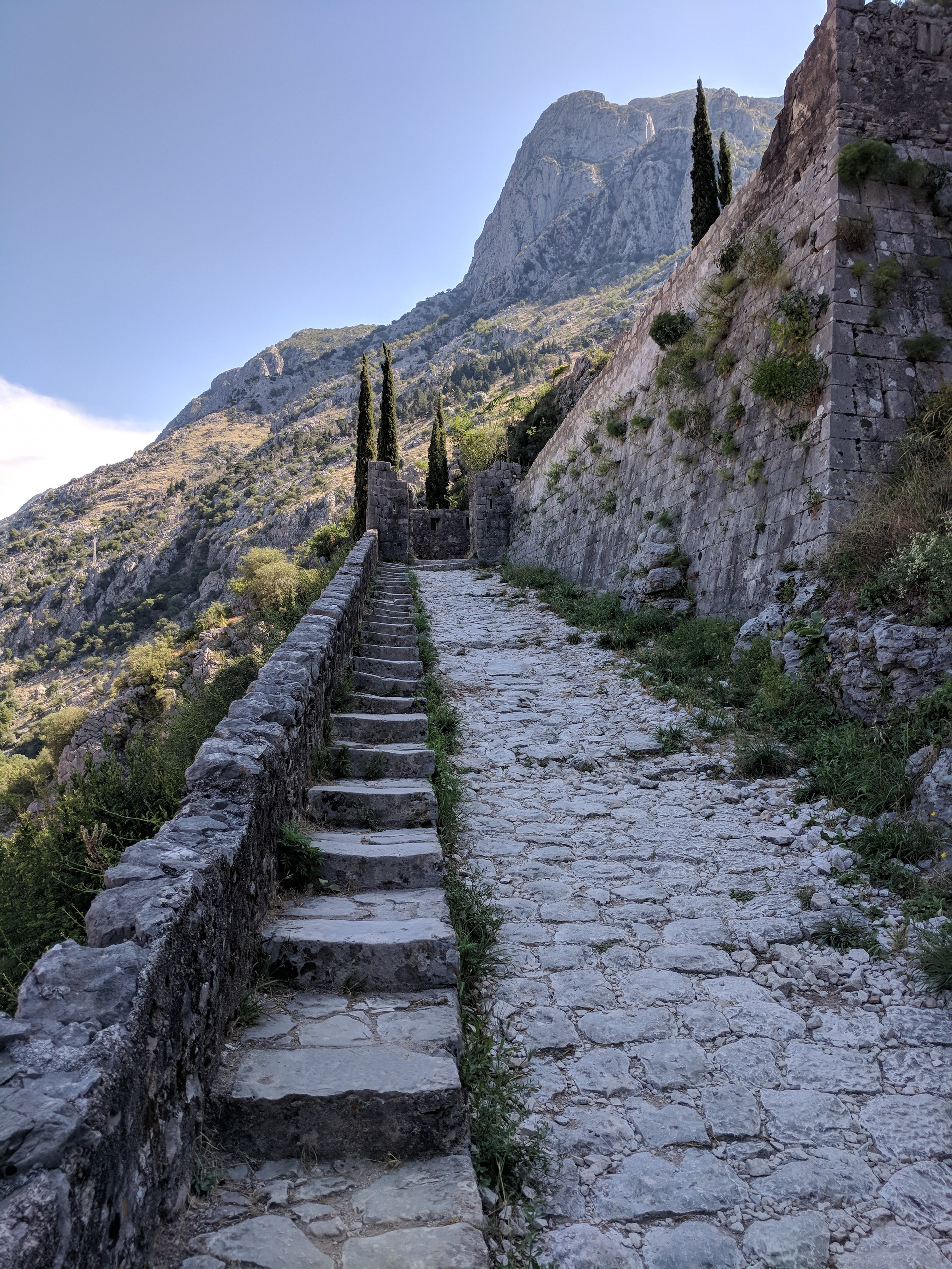 Cobblestone pathway with stone steps and cypress trees, flanked by a stone wall and mountain landscape under a blue sky.