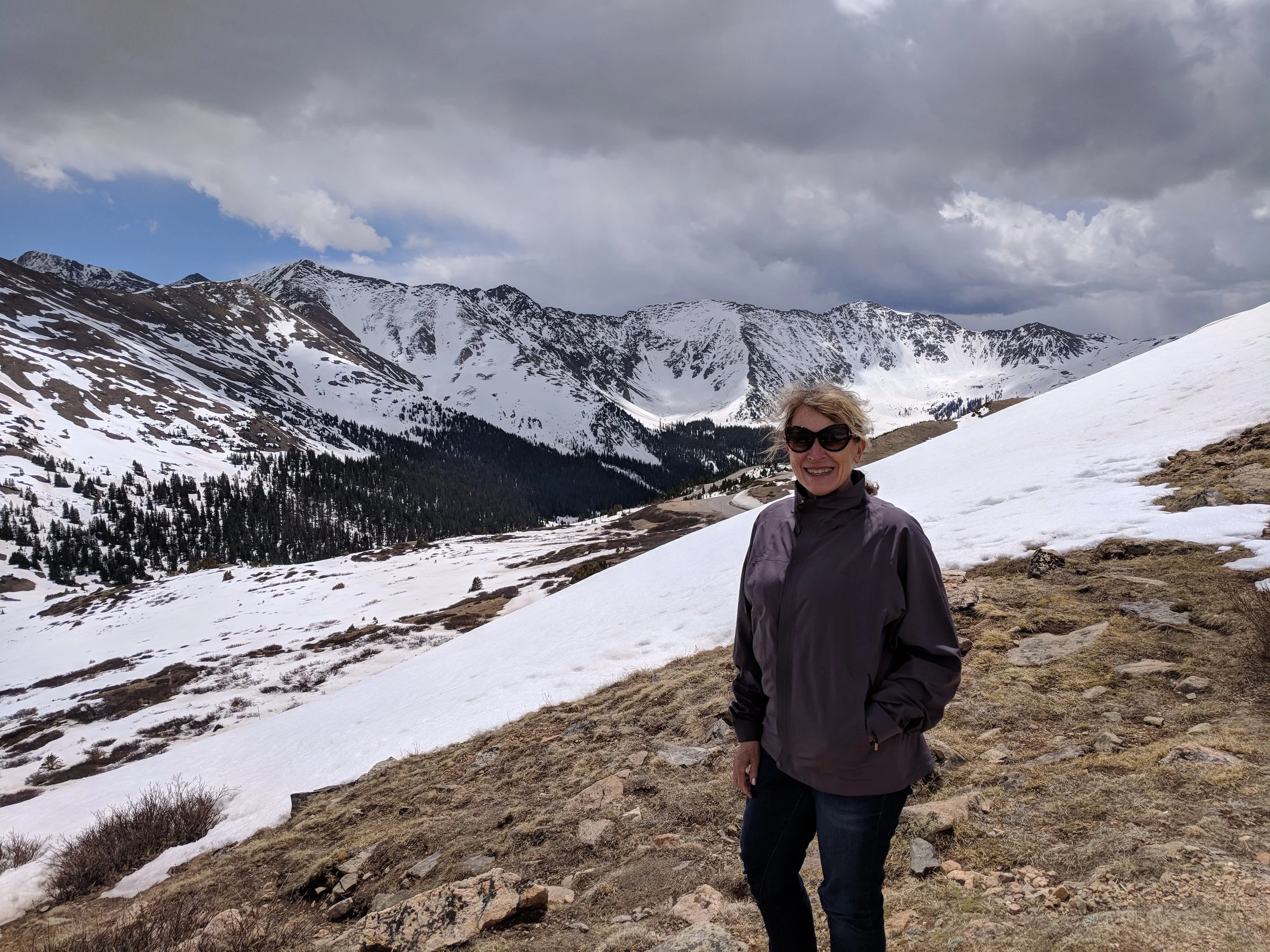 Woman in sunglasses standing on a snowy mountain slope with a scenic view of snow-covered peaks and a cloudy sky in the background.