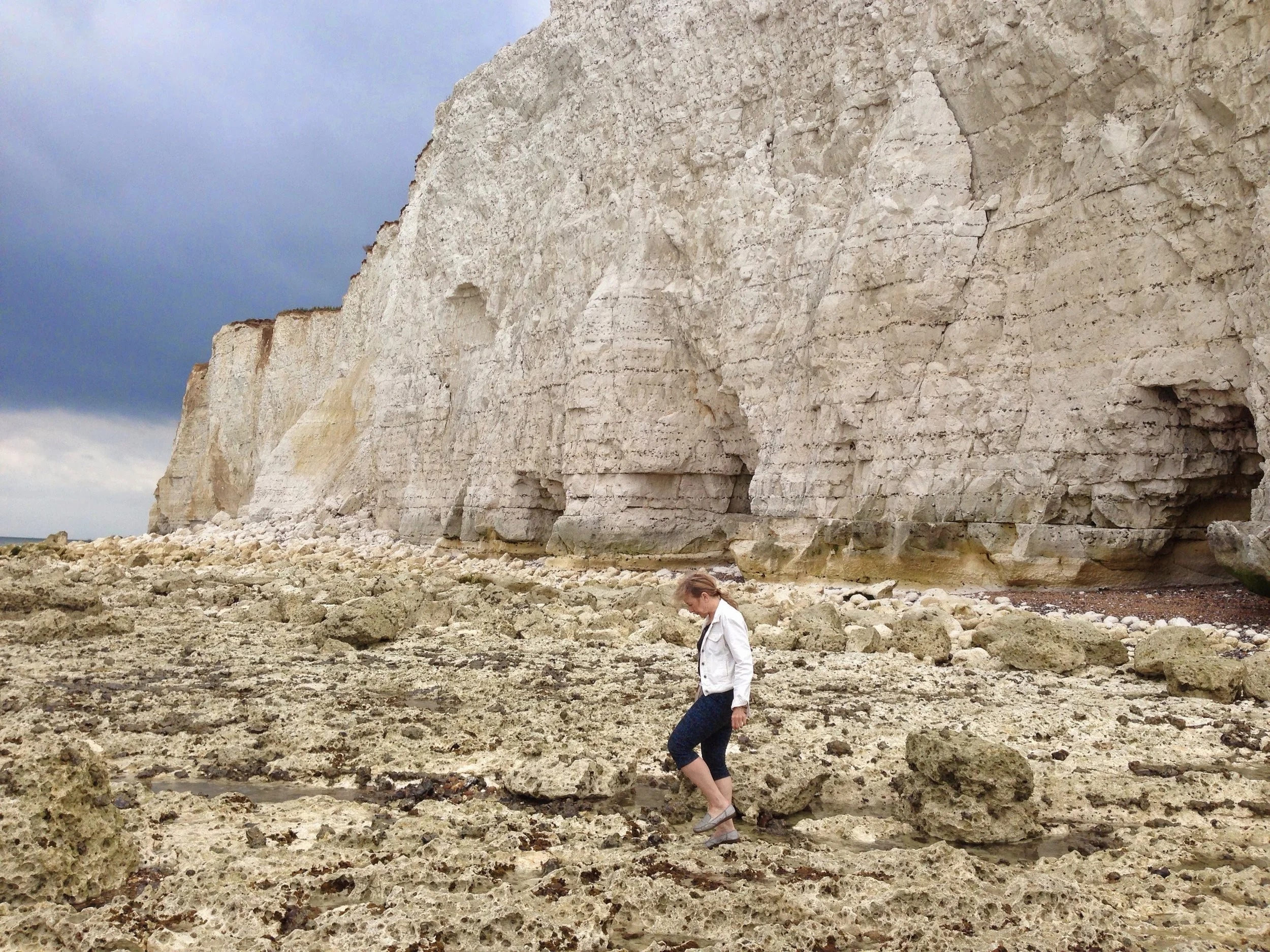 Person walking near white chalk cliffs on a rocky beach under a cloudy sky.