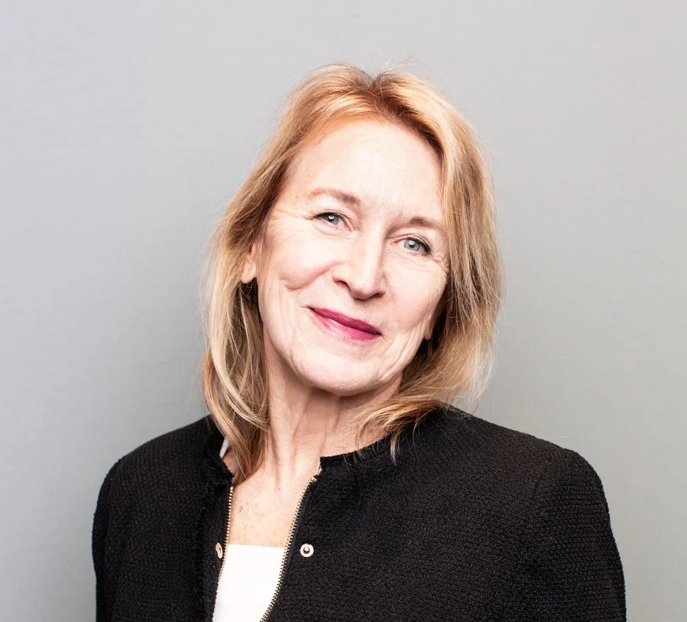 Portrait of a smiling woman with light-colored hair wearing a black jacket against a plain background.