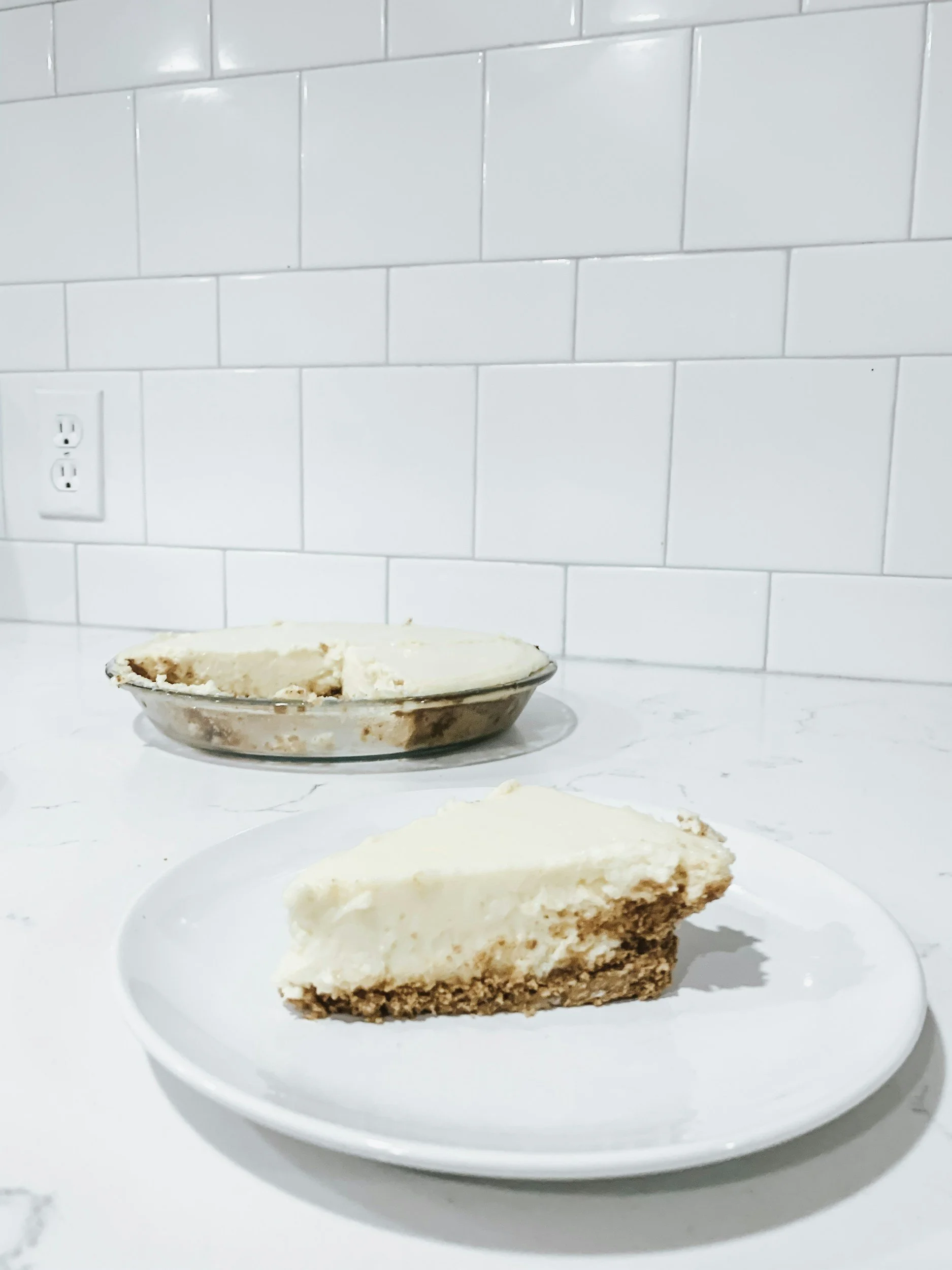 A slice of layered cake with cream frosting on a white plate, with a full cake in the background on a white surface, against a white tiled wall.