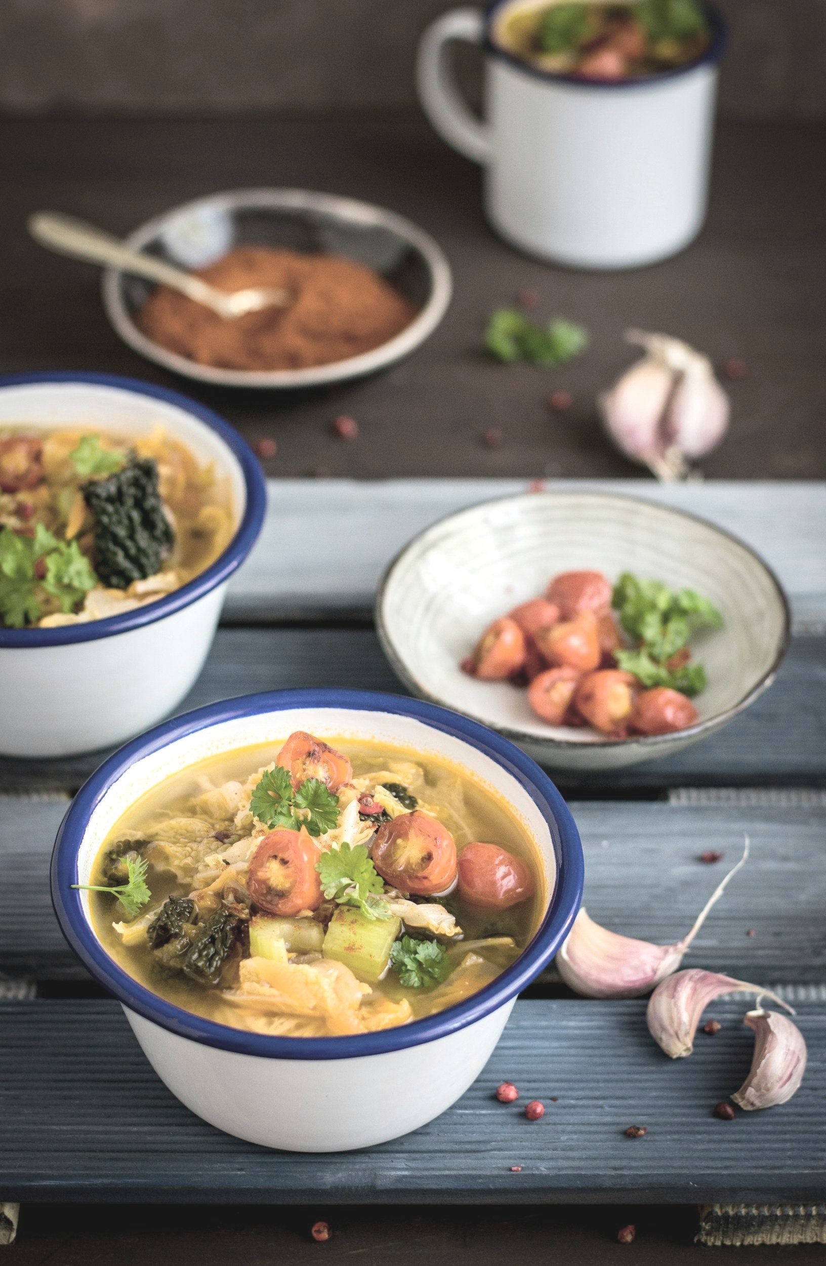 Bowls of soup with vegetables and herbs, garlic cloves, garlic powder, and peppercorns on a dark wooden surface.