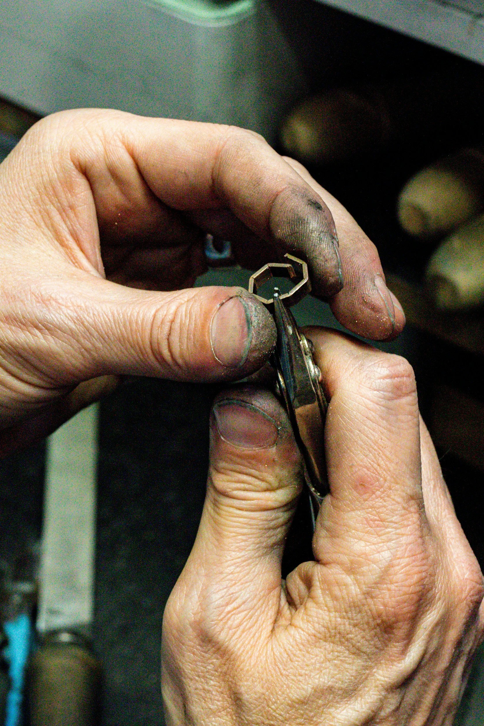 Close-up of a jeweller's hands assembling a small metal part, with visible work stains and dirt on the fingers.