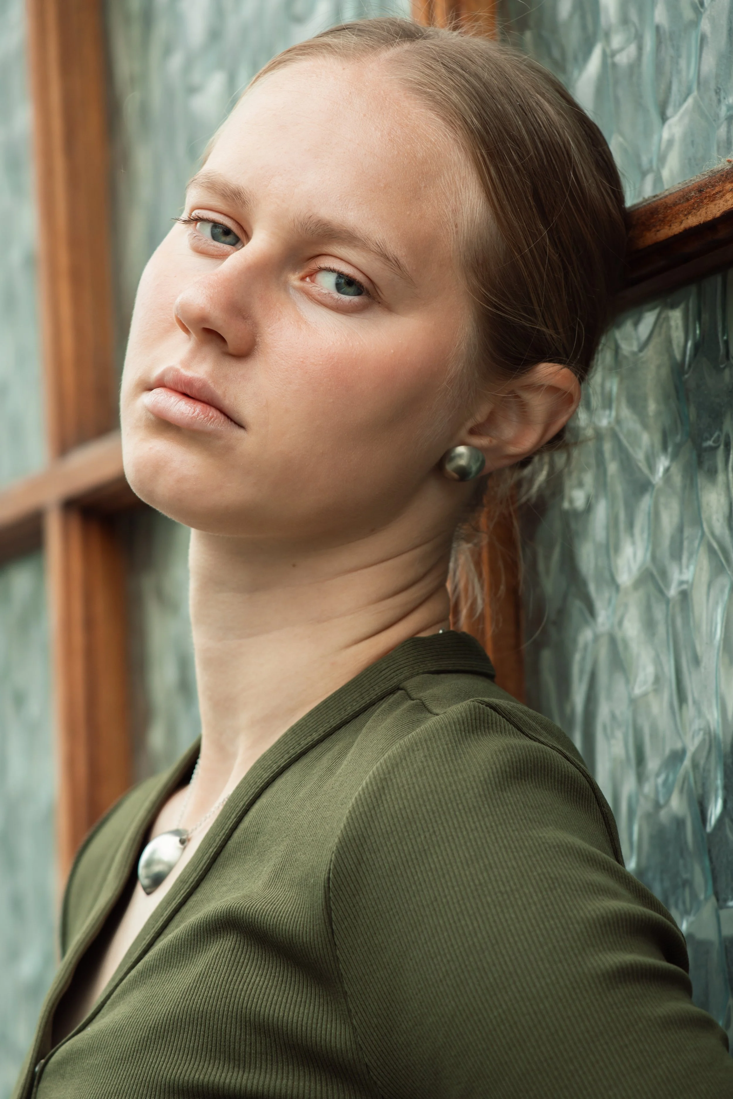 Close-up of a woman with light skin and blue eyes, leaning against a textured glass wall, wearing an olive green and Breners of London solid sterling silver Silvery Moon Earrings.