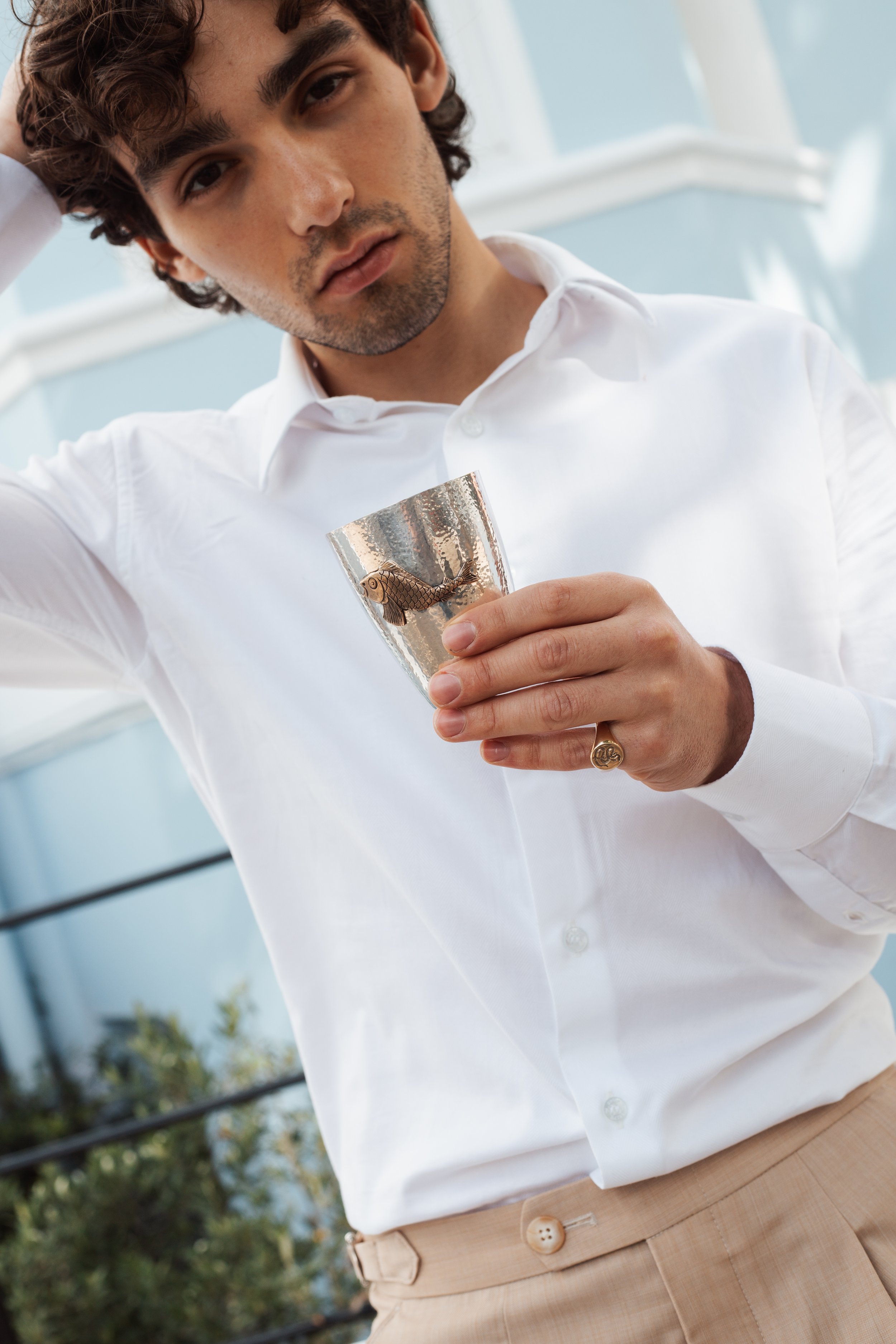 Young man with dark wavy hair, wearing a white dress shirt, holding a solid sterling silver beaker or cup, standing outdoors with blue sky and trees in the background.