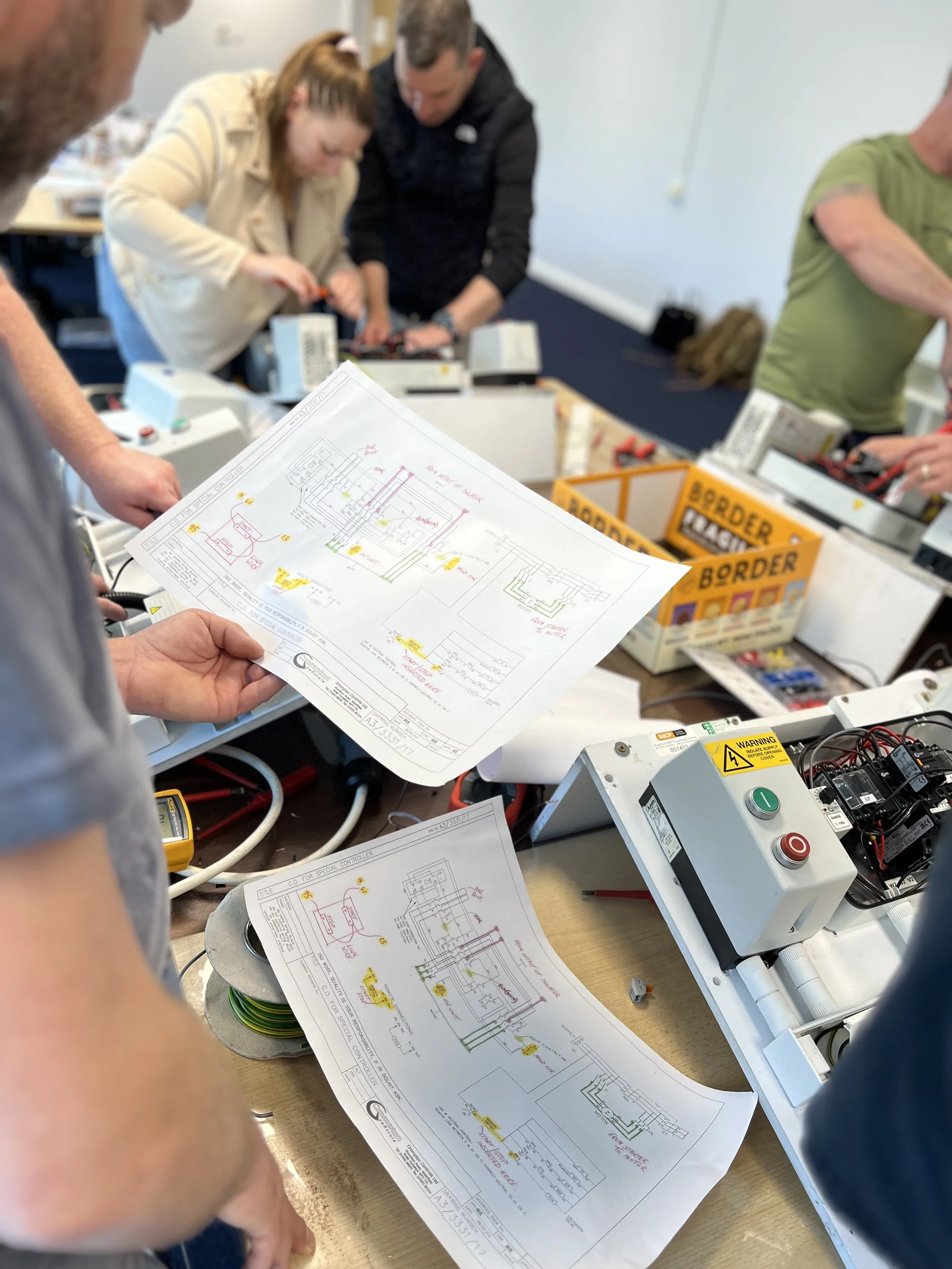 Group of people working on electrical engineering or wiring project with diagrams and equipment on a workshop table.