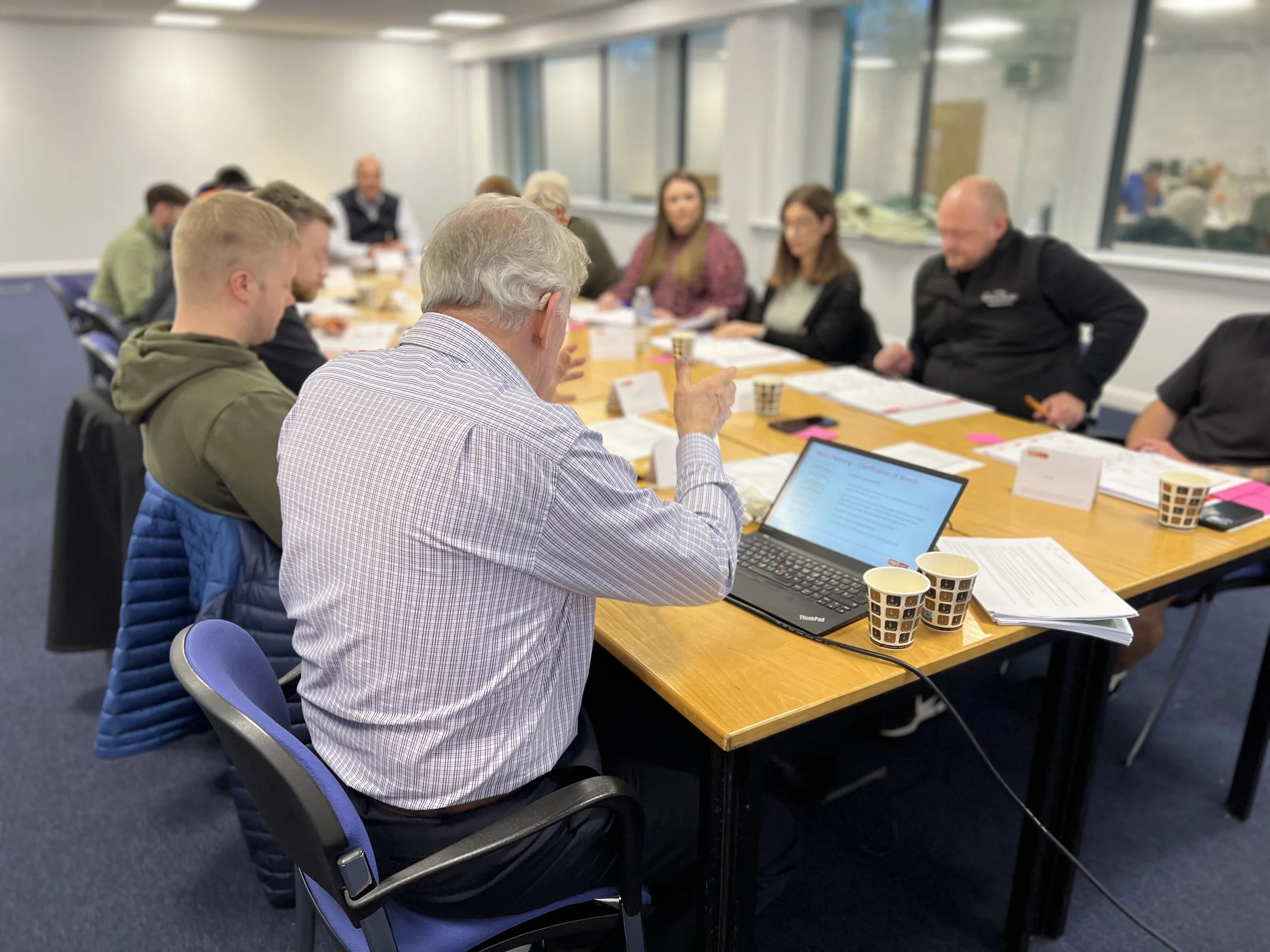 A group of people seated around a large conference table in a meeting room, with a man in the foreground using a laptop, and others listening.