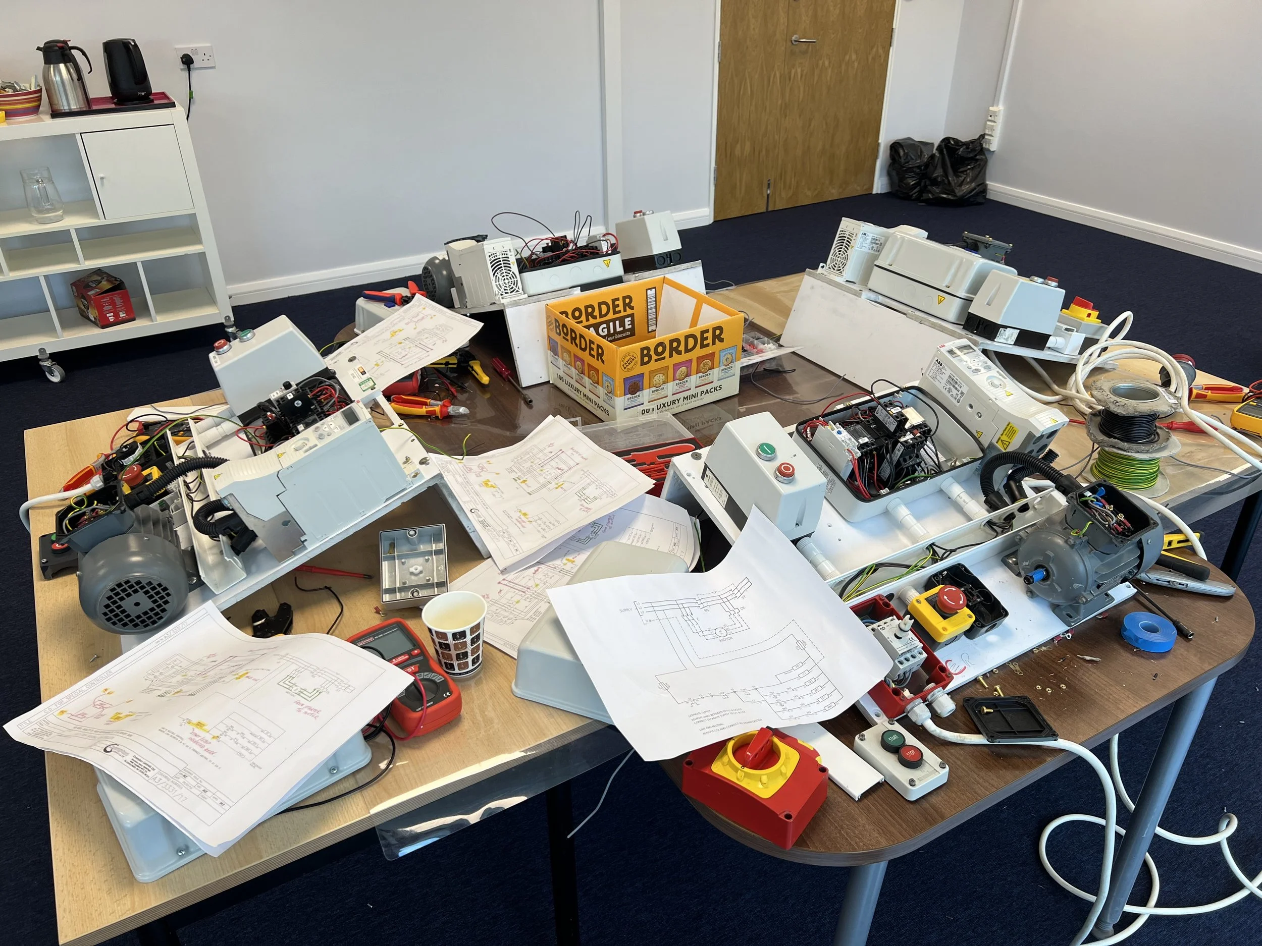 A worktable cluttered with electrical components, schematics, tools, and wires, likely for an electronics or engineering project.