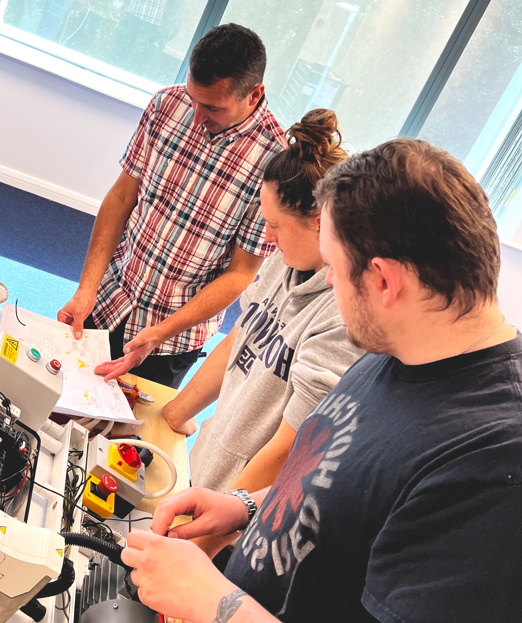 Four people gathered around a table with electrical equipment and diagrams, engaged in a hands-on training or workshop on electrical systems.