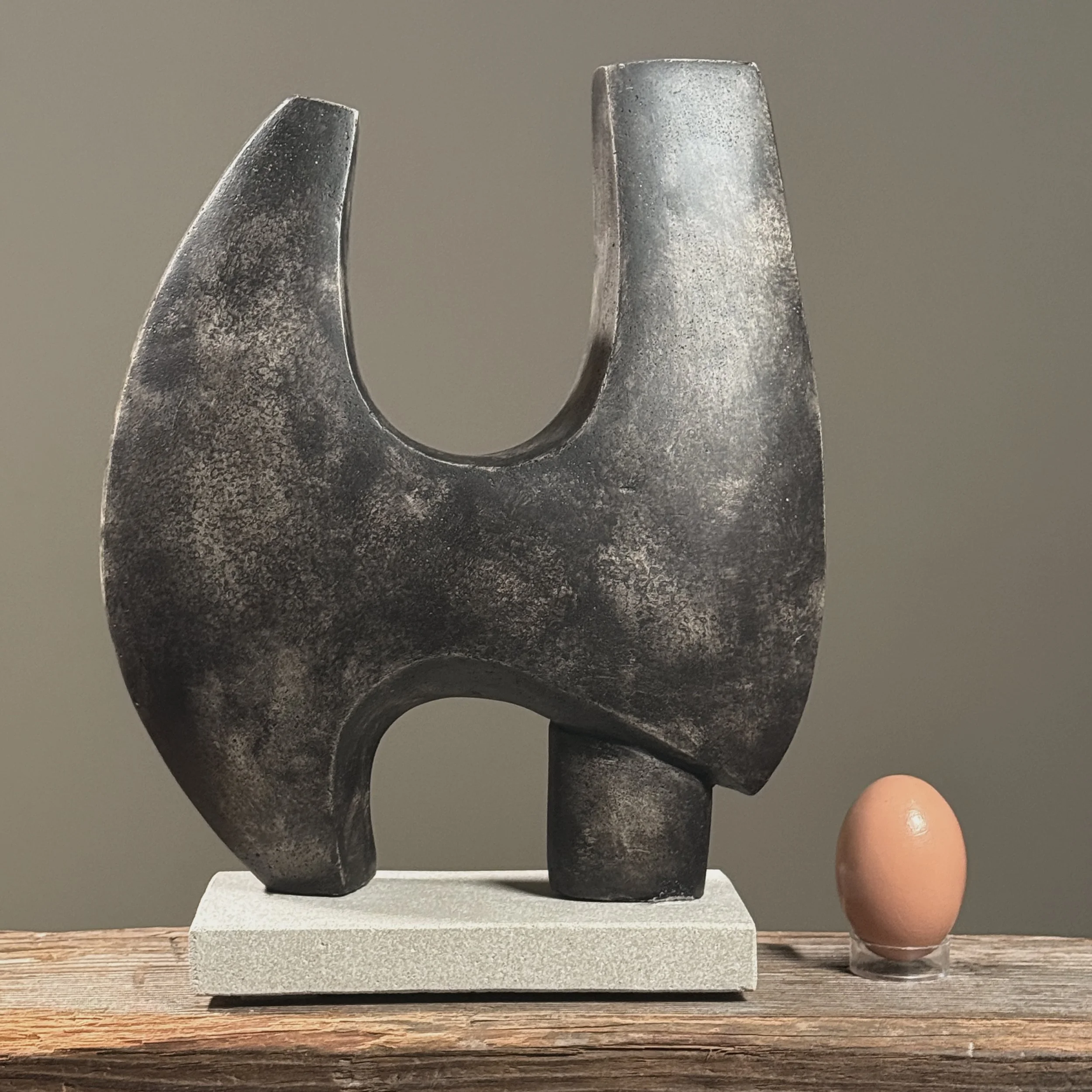Full frontal view of a modernist biomorphic cement sculpture with dark grey mottled patina and winged form With egg for size reference.