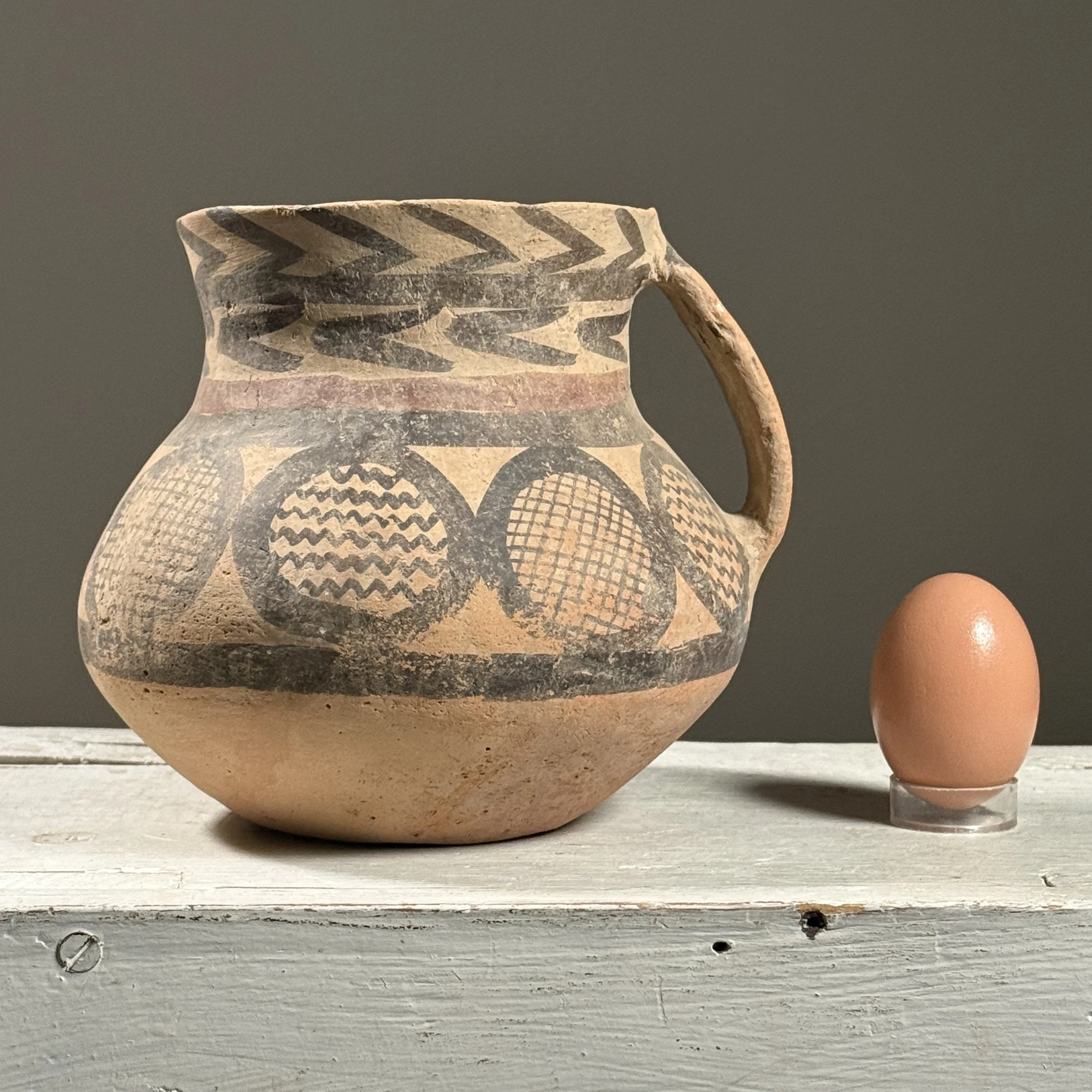 Full frontal view of a Chinese Neolithic style pottery jar with bulbous body and flaring neck with an egg for size reference.