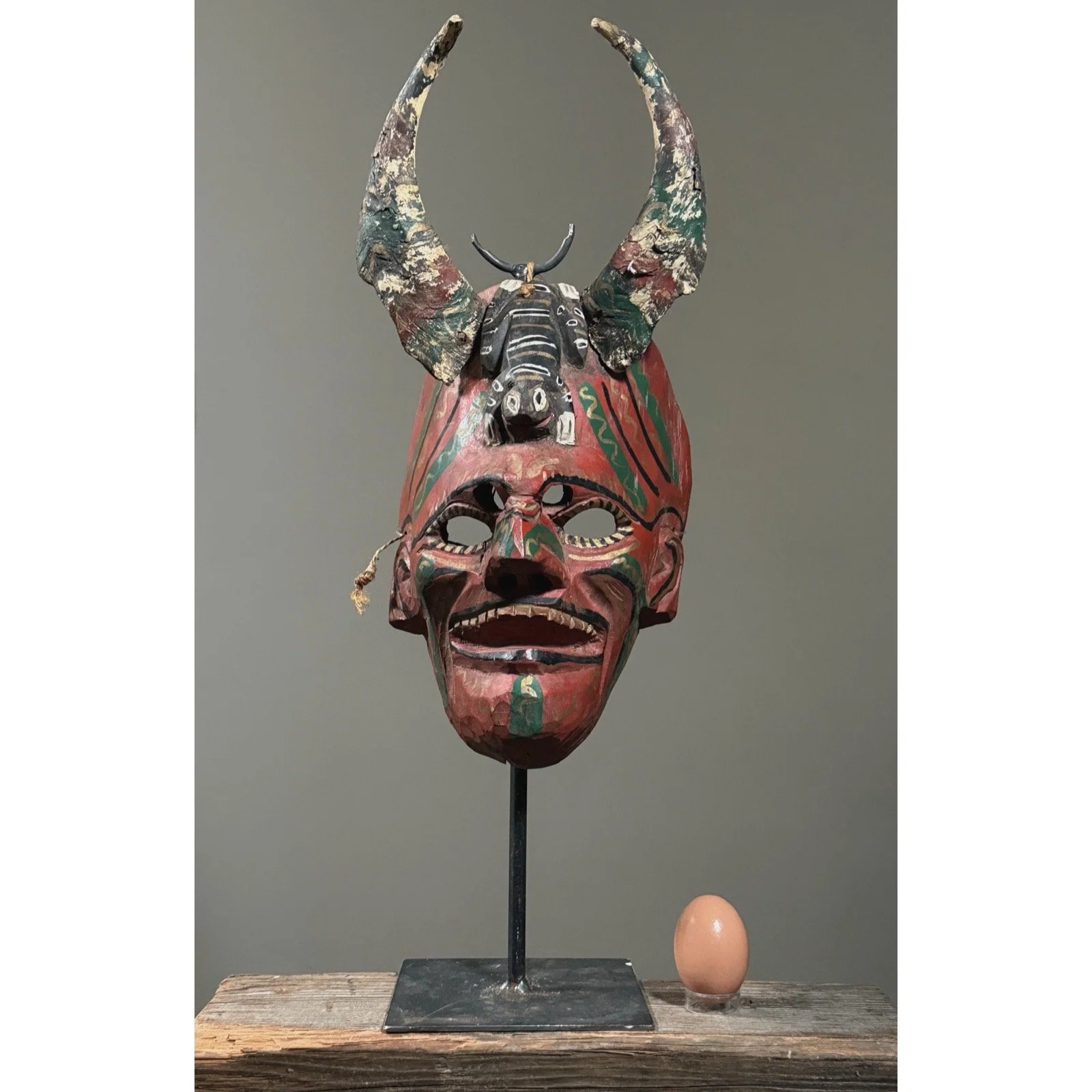 Front view of a vintage Mexican carved devil mask with horns with an egg for size reference.