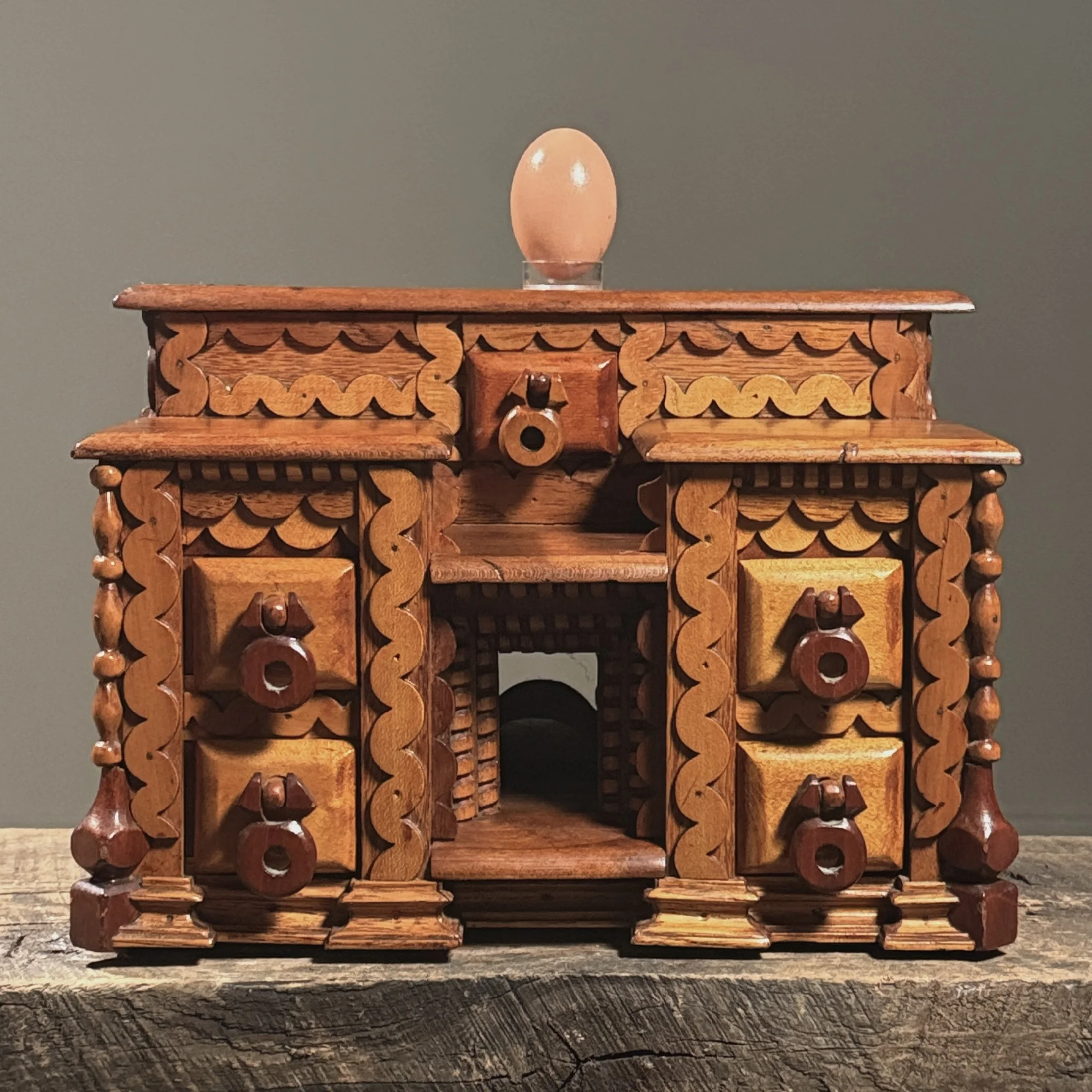 Full frontal view of a vintage folk art box with seven drawers and heavy layered scrollwork with egg for size reference.