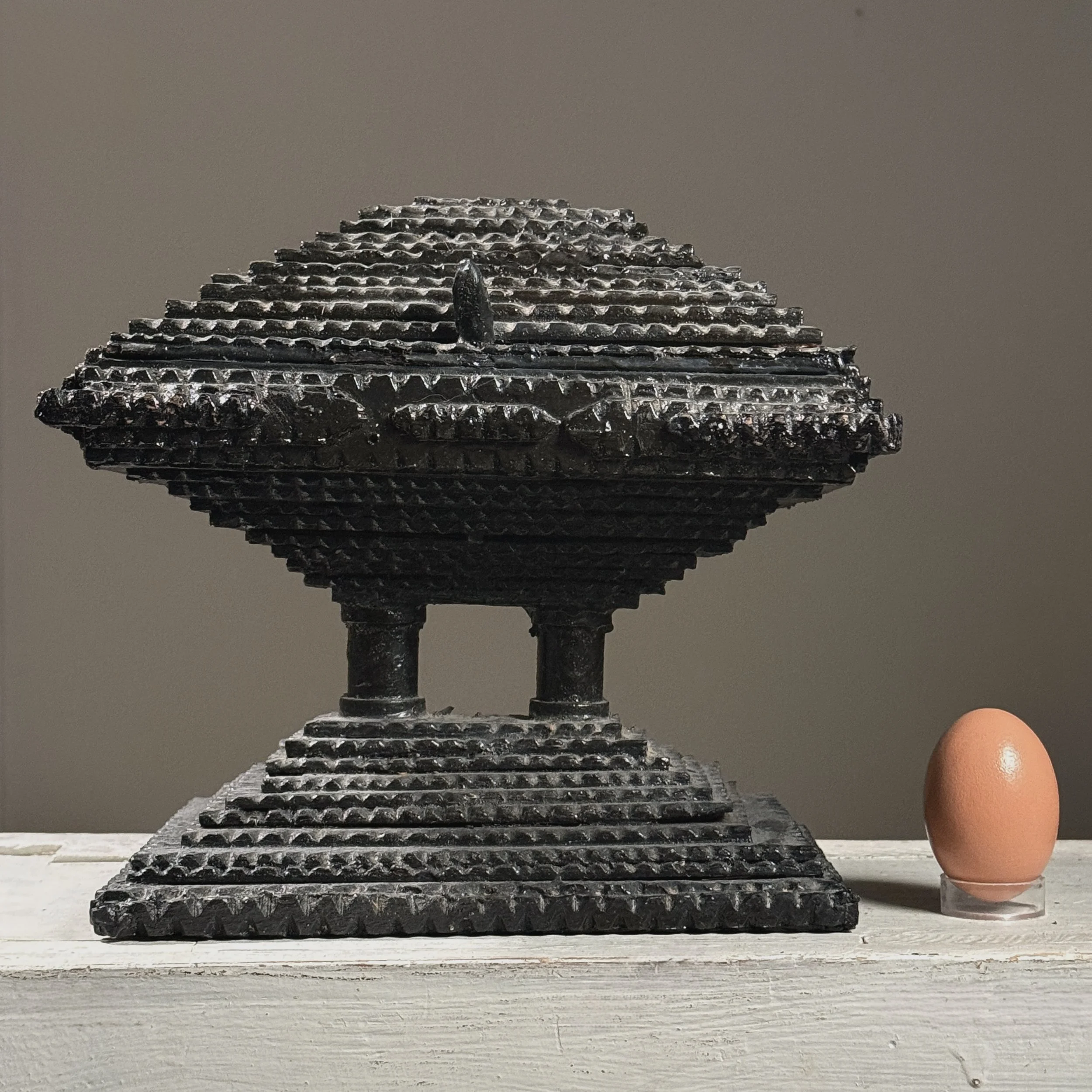 Full frontal view of an antique black painted chip-carved tiered tramp art box with hinged top with egg for size reference.