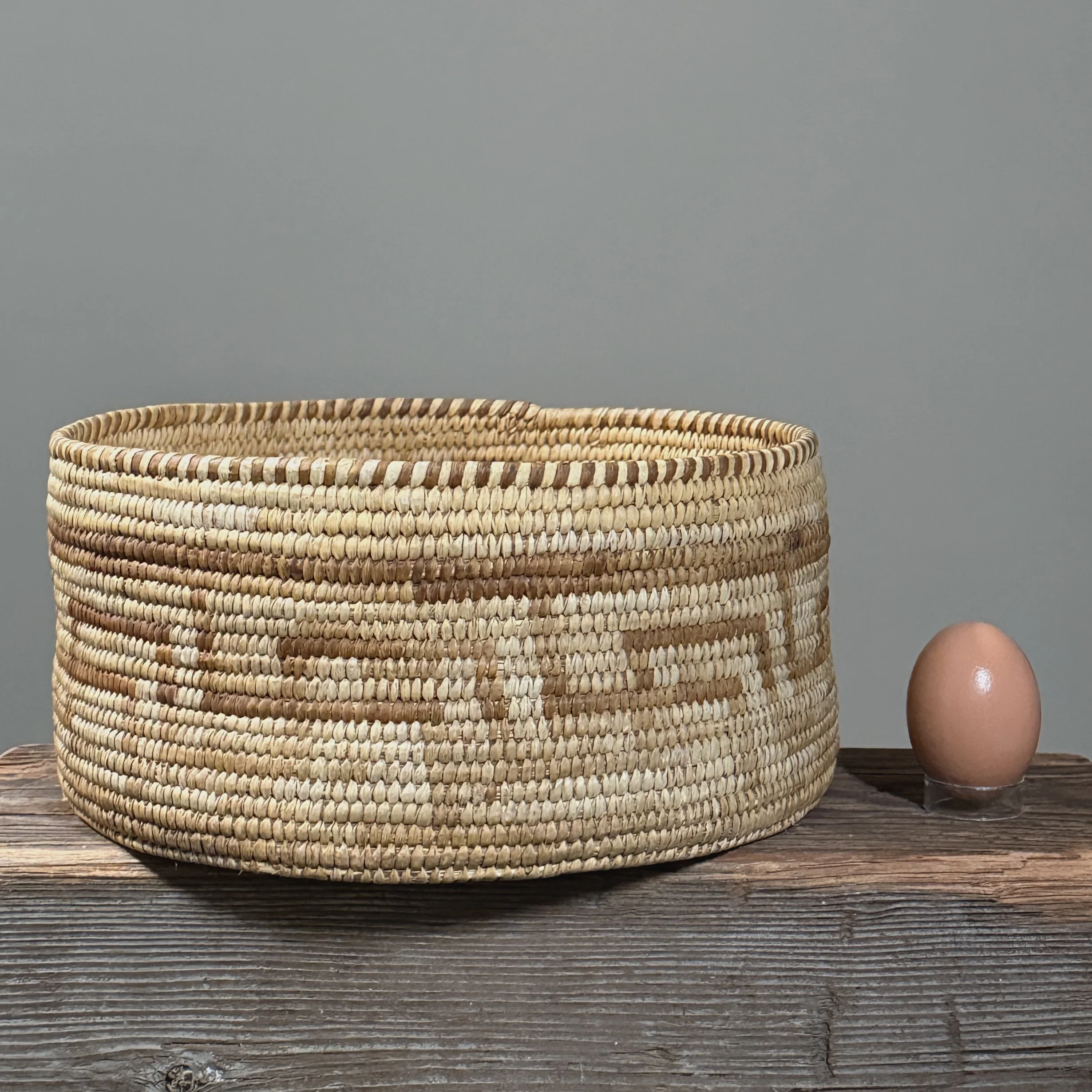 Front view of an antique Pima coiled basket with high straight walls with egg for size reference.