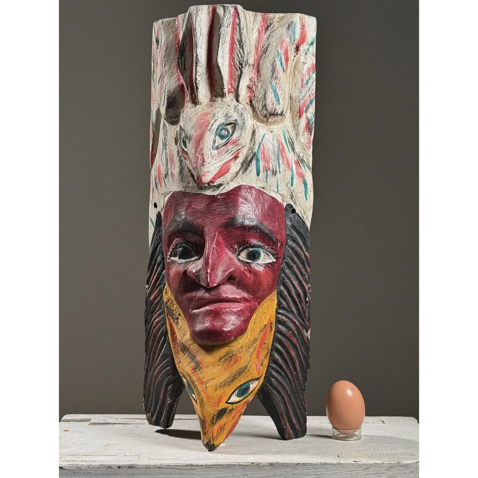 Front view of a vintage Mexican folk art mask with red-painted human face with an egg for size reference.