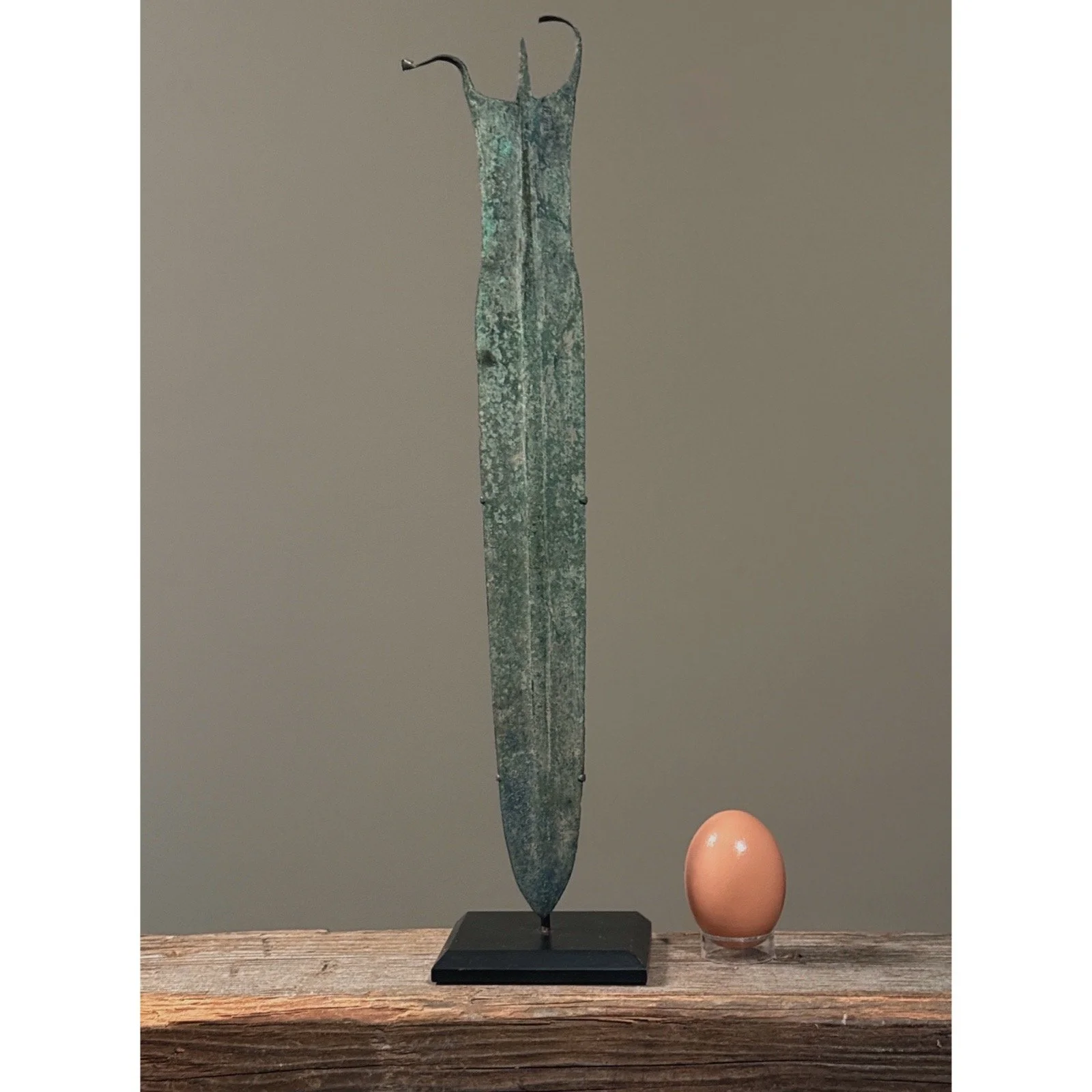 An ancient Luristan bronze dagger mounted on a custom metal display stand with an egg for size reference.