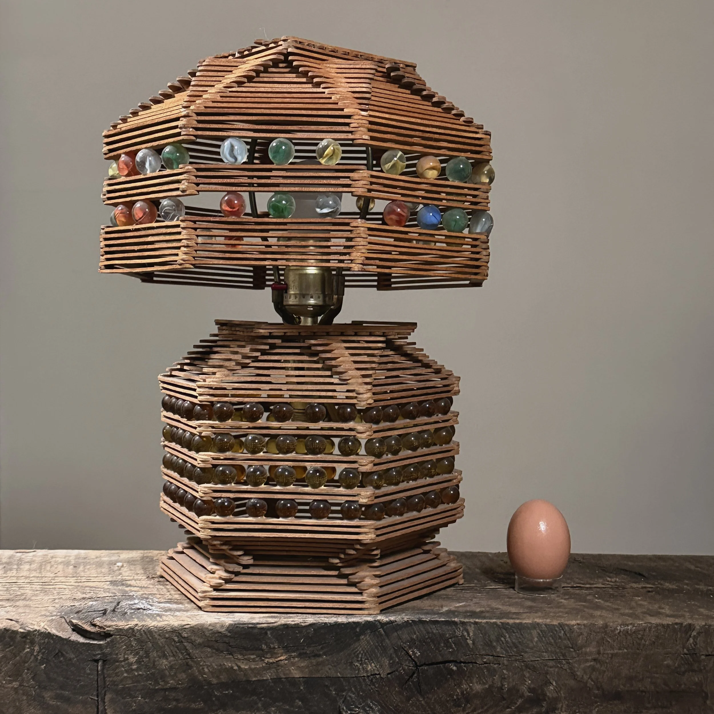 Full front view of the functional vintage popsicle stick and marble folk art table lamp with an egg for size reference.